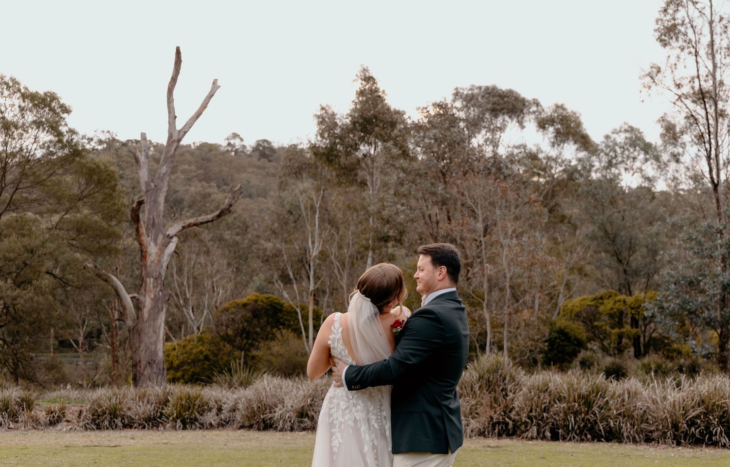 A couple walks together in a lush outdoor garden setting with tall trees in the background.