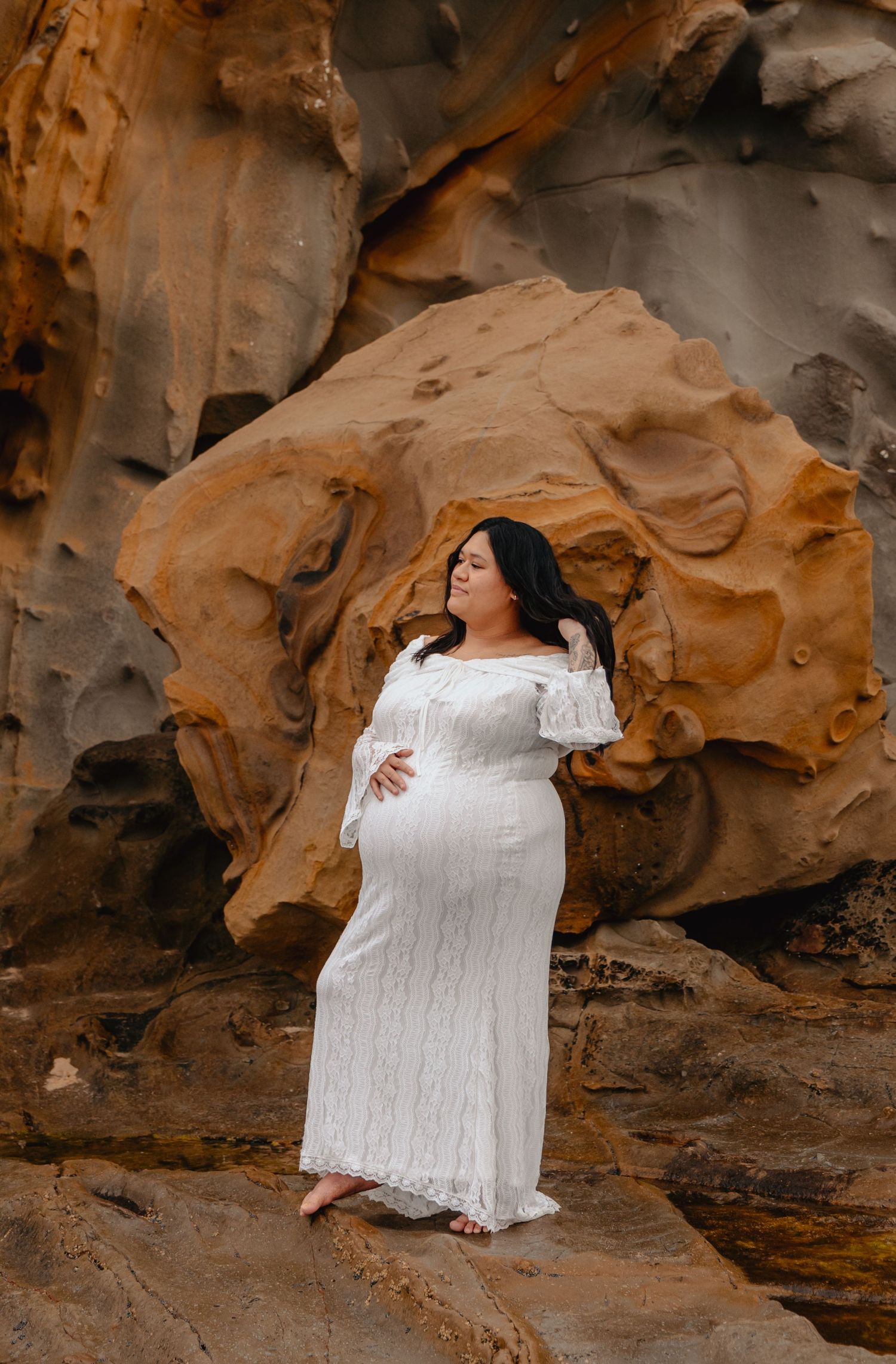 A figure in a flowing white dress poses gracefully against textured sandstone rock formations in warm golden light.