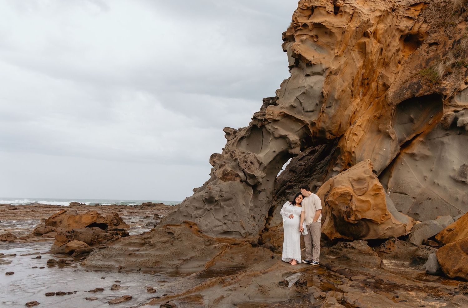 A couple in white attire poses together on a rocky beach during sunset, creating a romantic coastal scene.