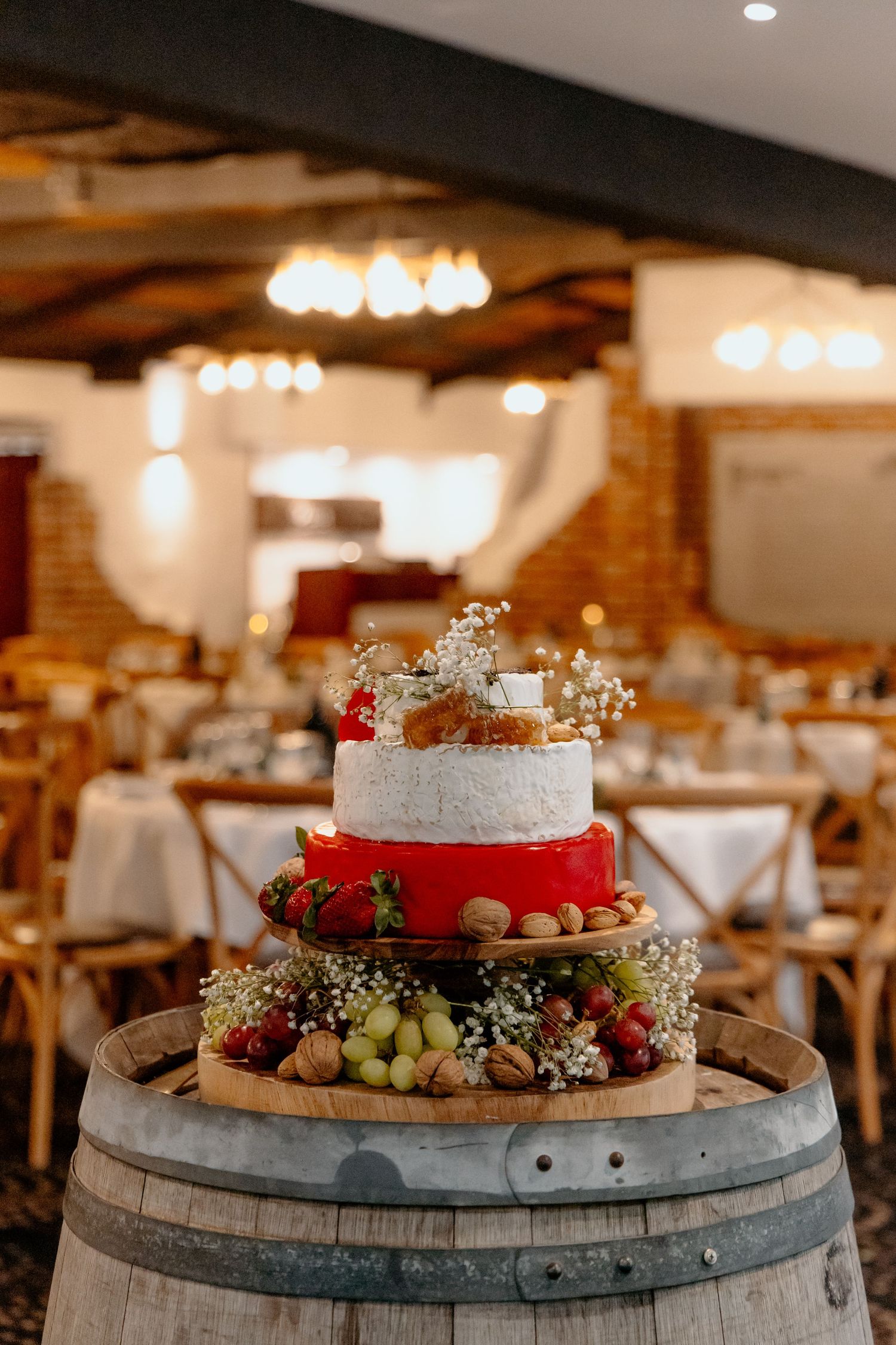 A rustic wedding cake decorated with red ribbon and fresh fruit sits atop a wooden wine barrel in a warm-lit reception venue.