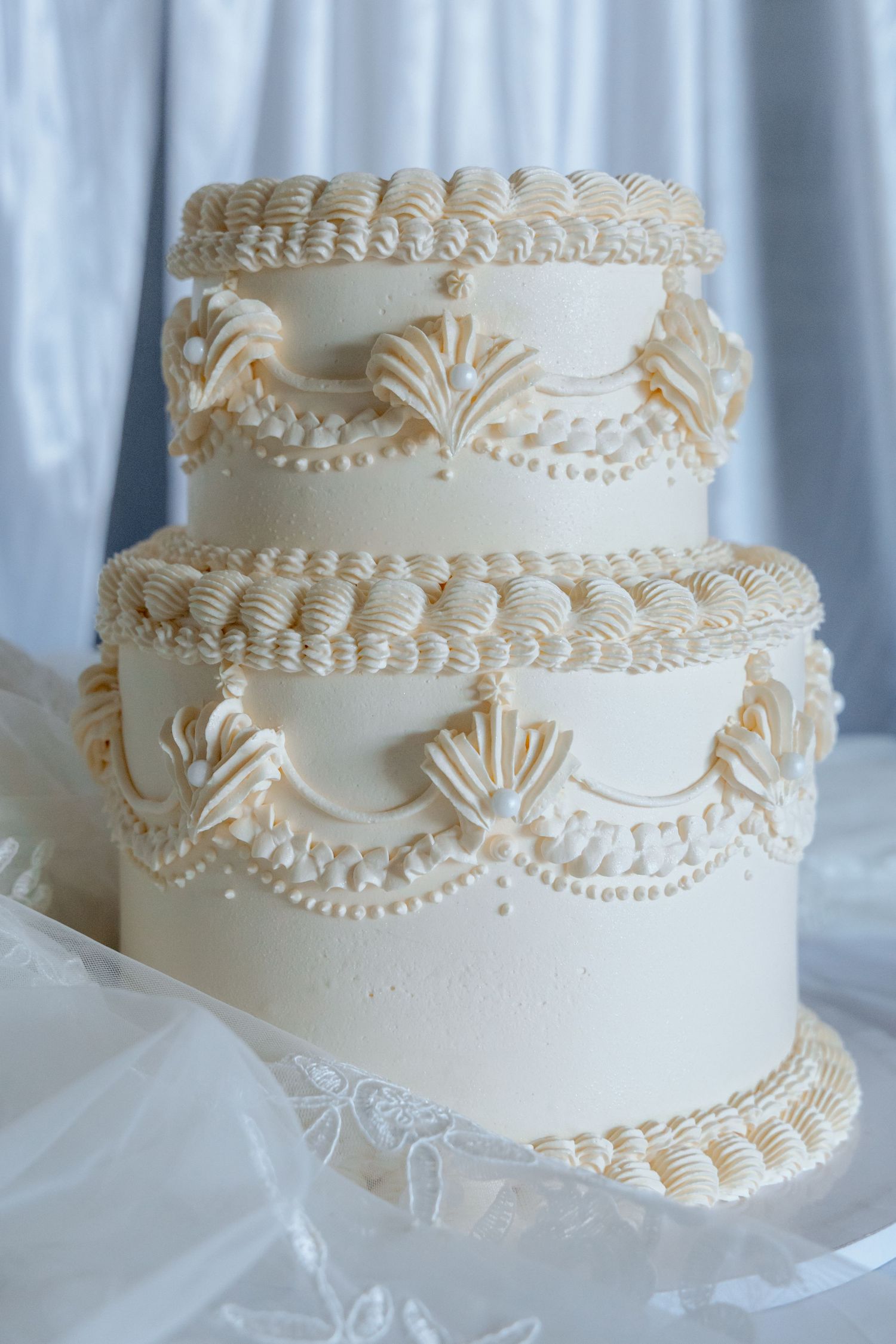 Elegant two-tier white wedding cake with intricate piped frosting details and shell decorations on a lace base.