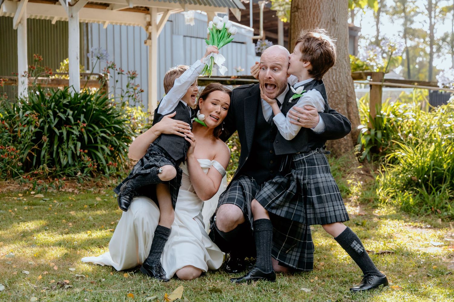 Wedding couple in kilts laugh with two children in a fun outdoor group photo on grass.