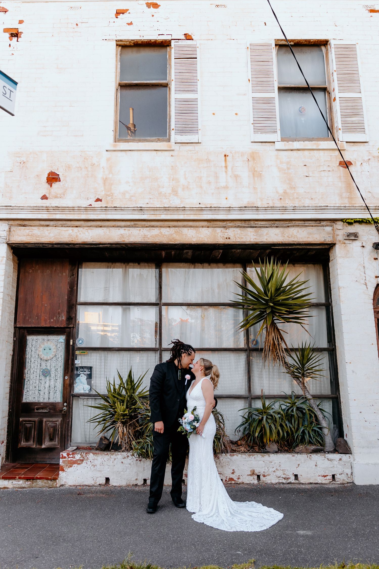 Bride and groom share a kiss in front of a rustic white building with vintage windows.