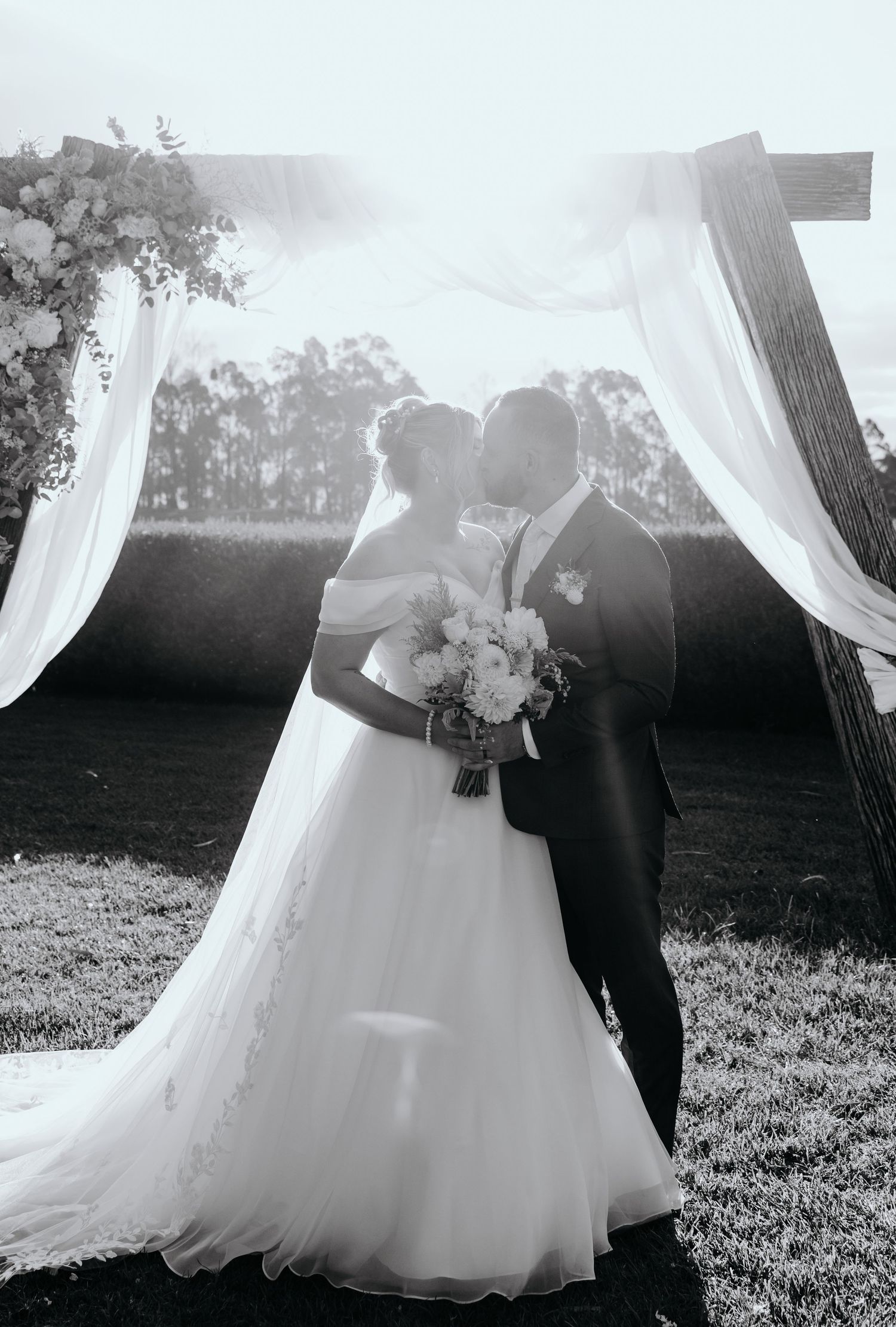 Black and white photo of a bride and groom kissing under a draped wooden arch with floral arrangements at an outdoor wedding.