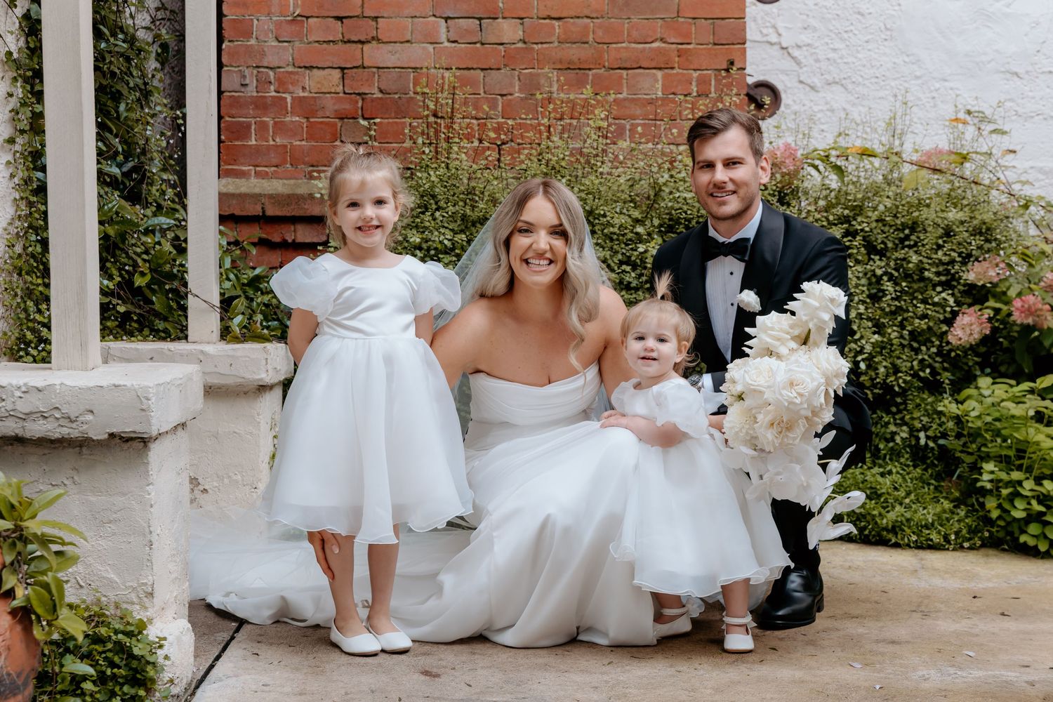 Bride and groom pose with two young flower girls in white dresses outside a brick venue on their wedding day.