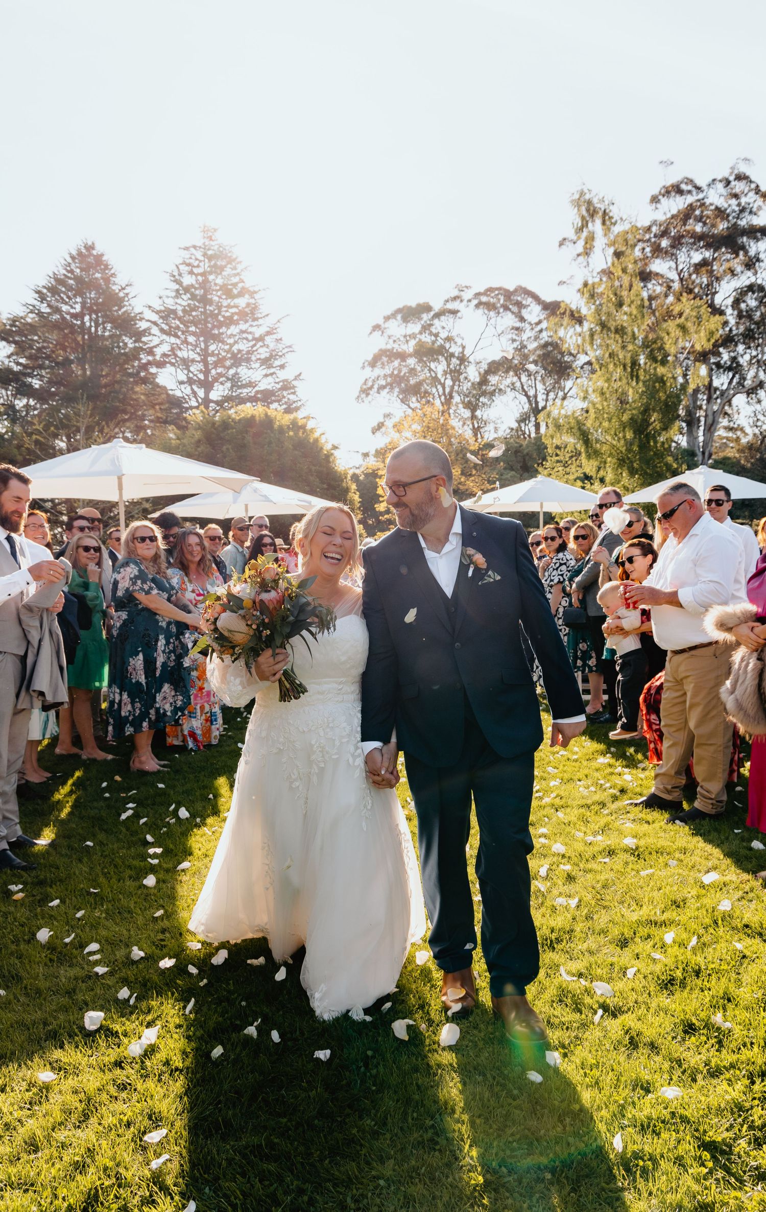 Wedding guests throw white flower petals as newlyweds walk through on a sunny grassy lawn during an outdoor celebration.
