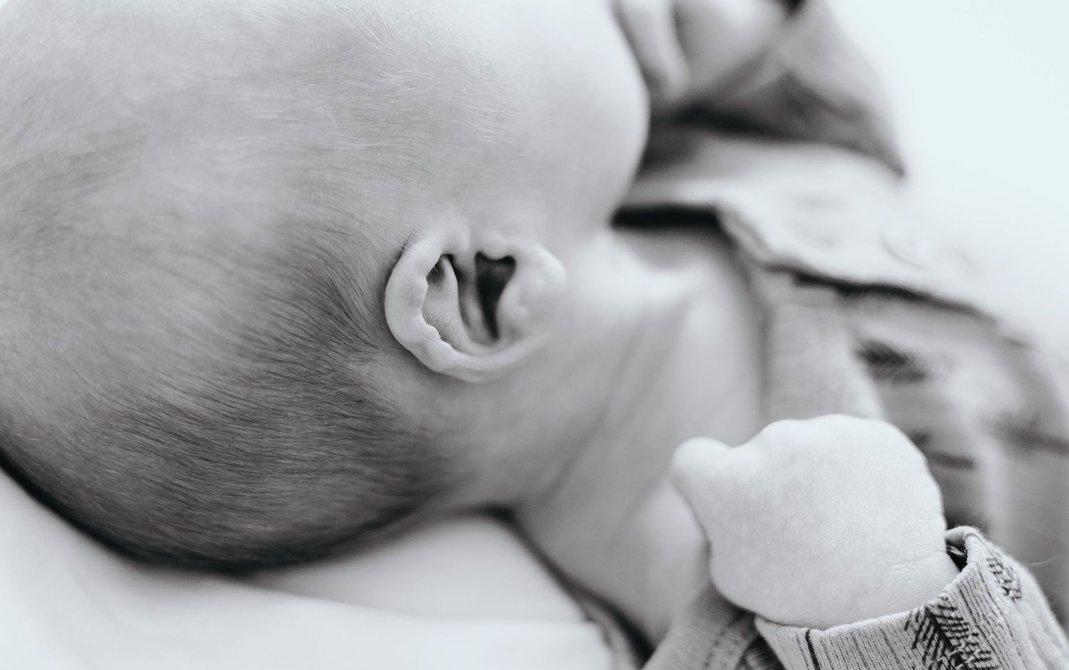 Close-up black and white photo of a sleeping baby's ear and head against soft bedding.