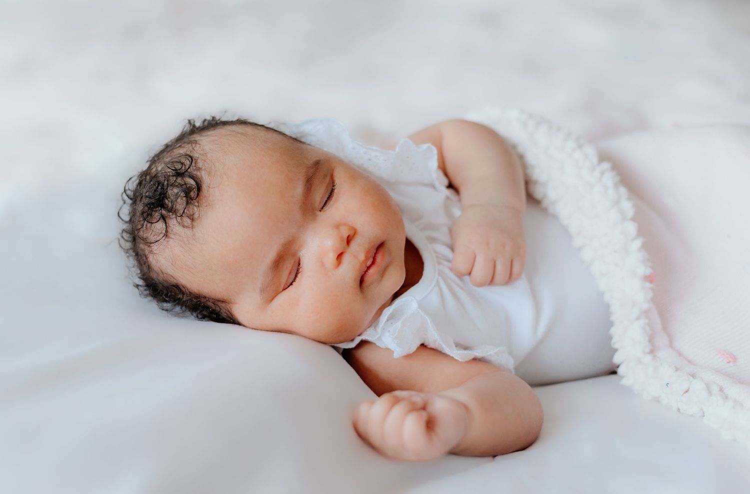 A sleeping newborn baby wrapped in soft white blankets lies peacefully against a light backdrop.