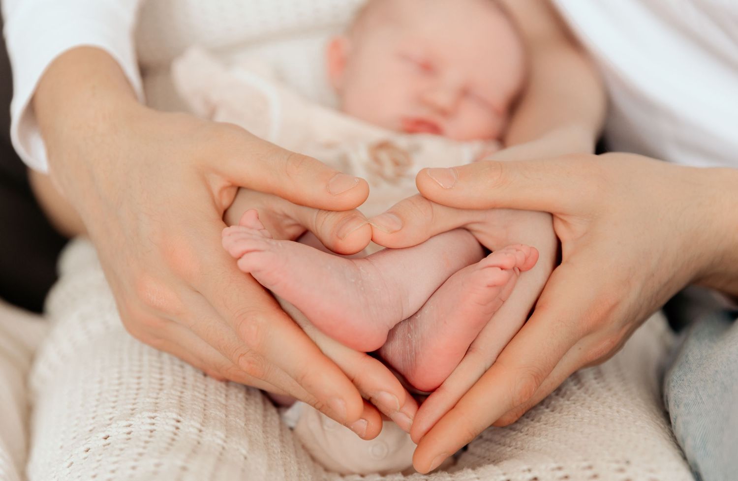 Close up of hands forming heart shape around baby's feet wrapped in white blanket.