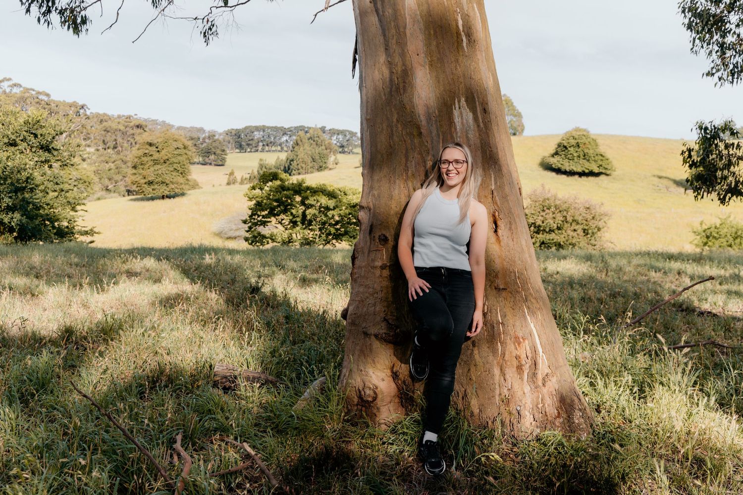 Person leaning against large eucalyptus tree trunk in sunny rural landscape with rolling hills.