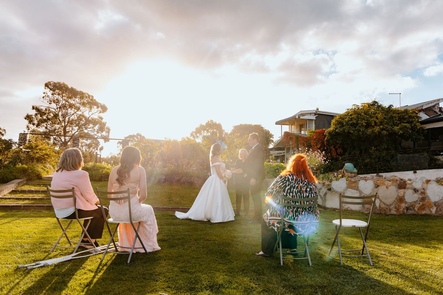 Small outdoor wedding ceremony on green lawn at golden hour with bride, groom, and seated guests.
