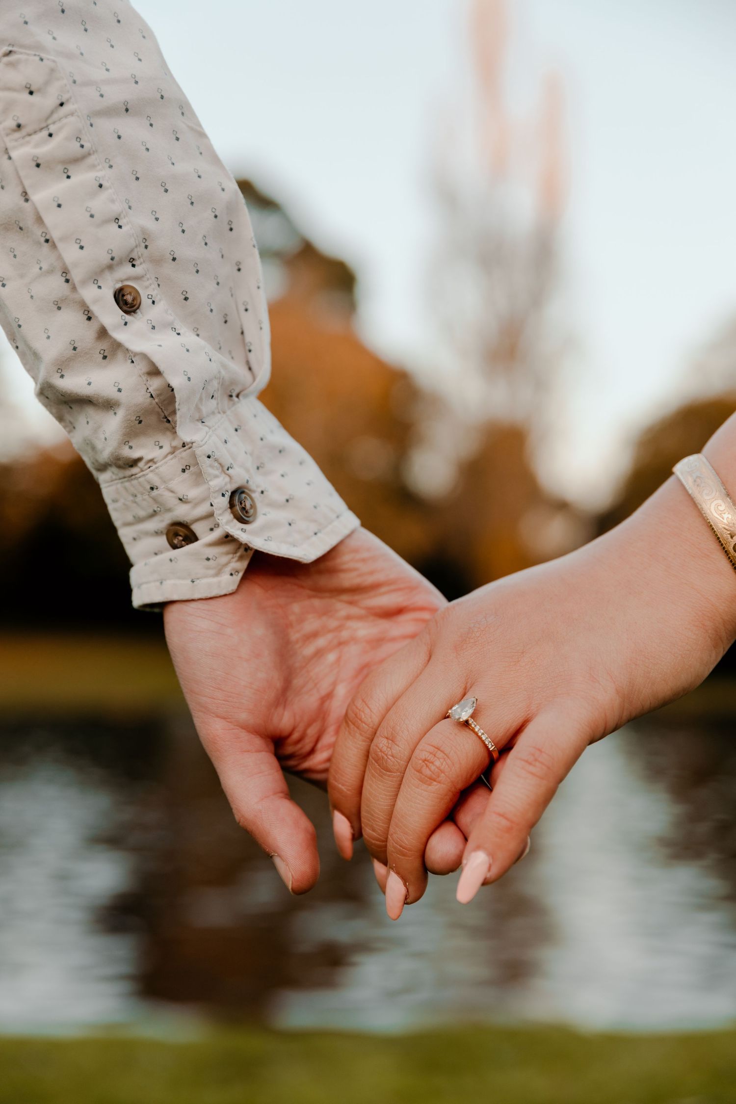 Close-up of two hands clasped together showing an engagement ring against a natural backdrop.