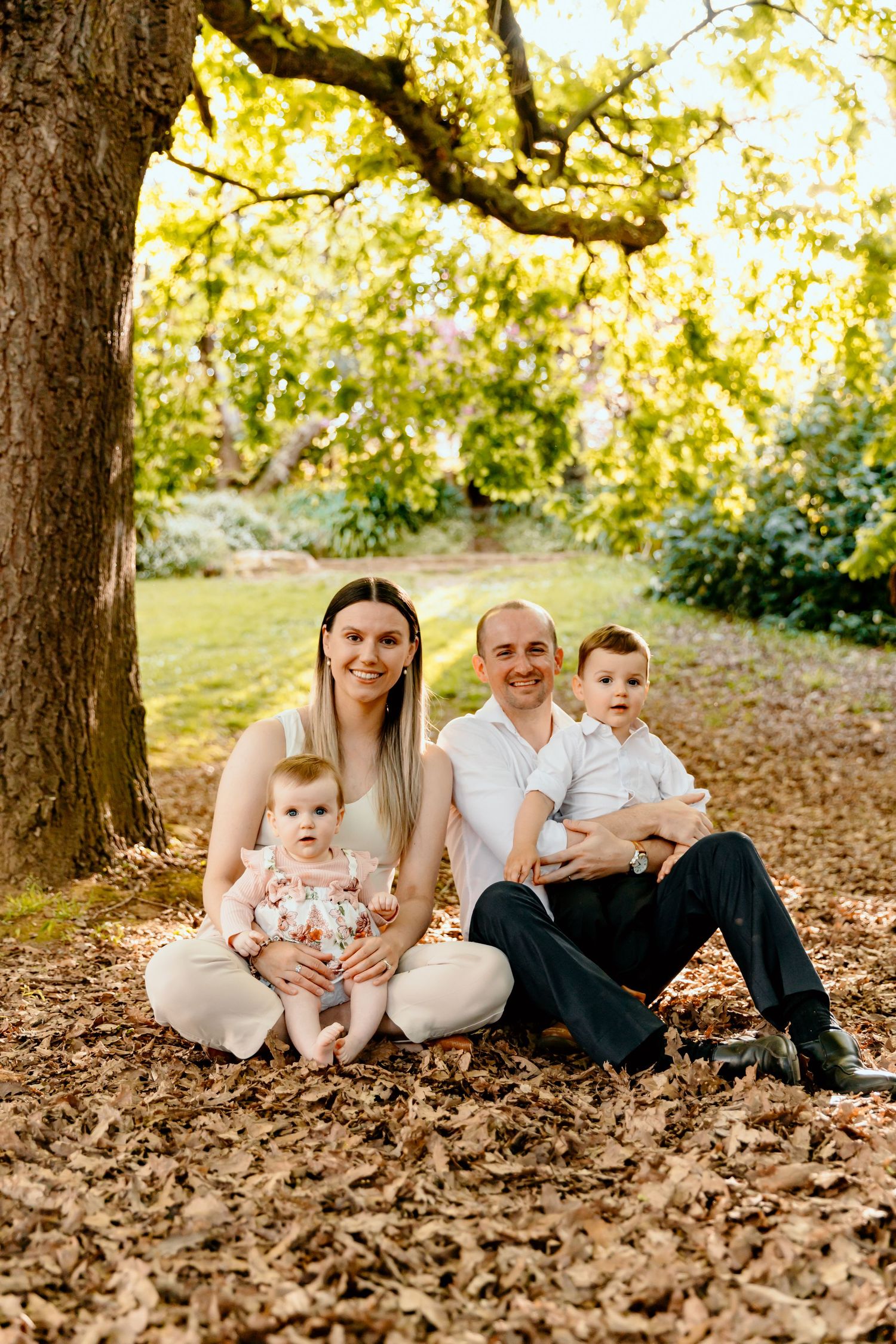 Family sitting together on autumn leaves under large tree in park.