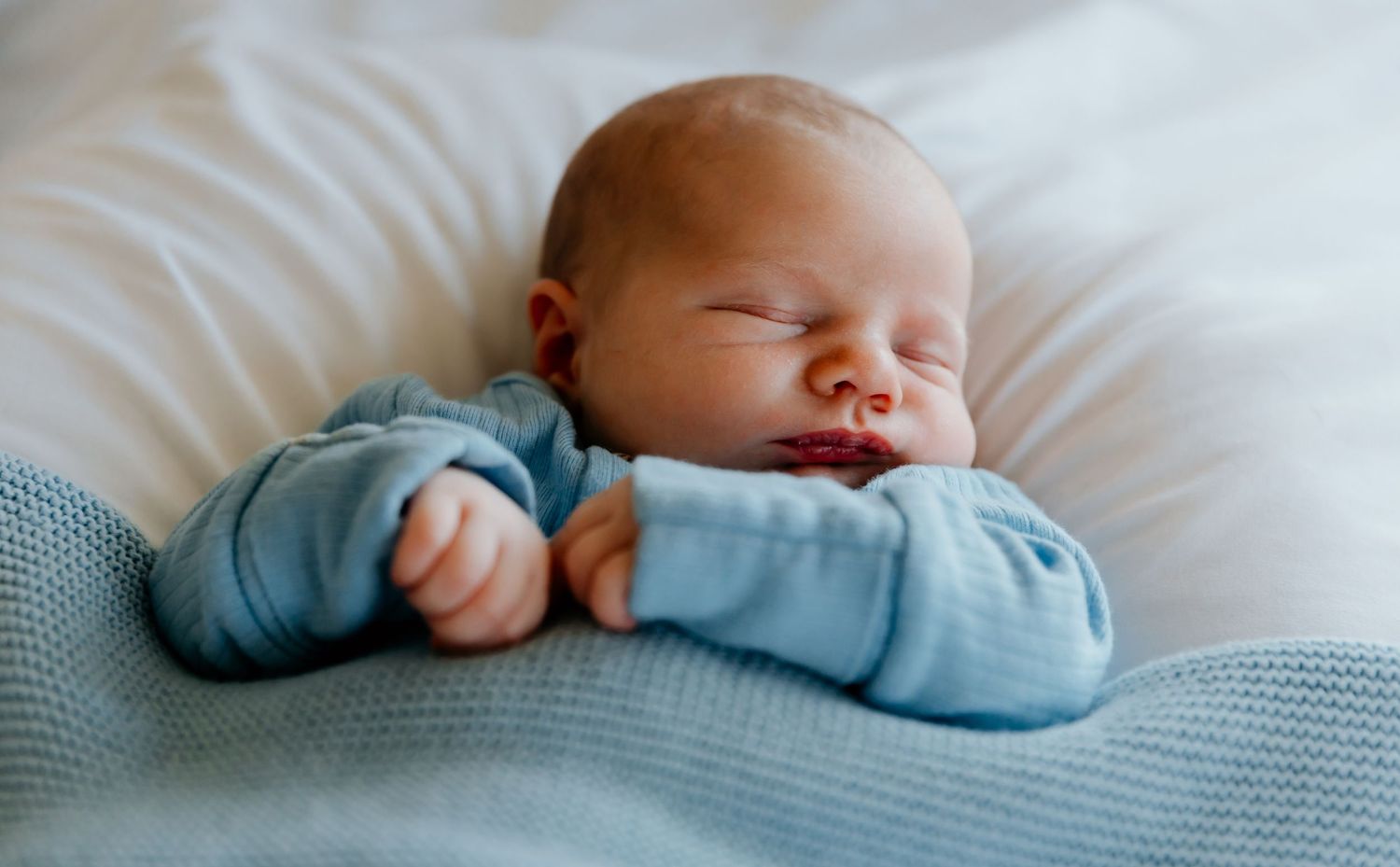 Peaceful sleeping infant wearing light blue outfit on white bedding.