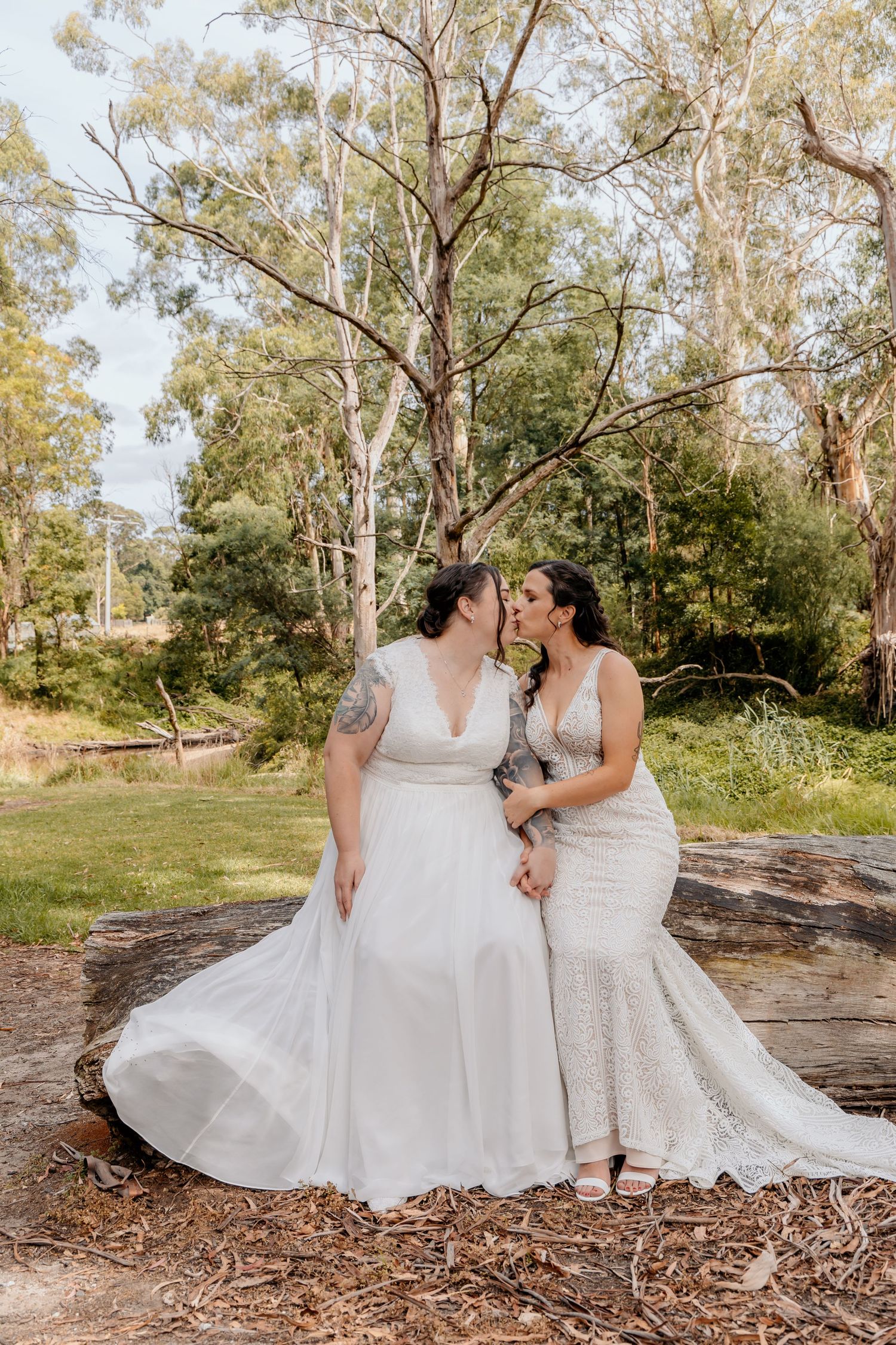 Two brides in elegant white wedding dresses pose together on a fallen tree trunk in a sunny outdoor forest setting.
