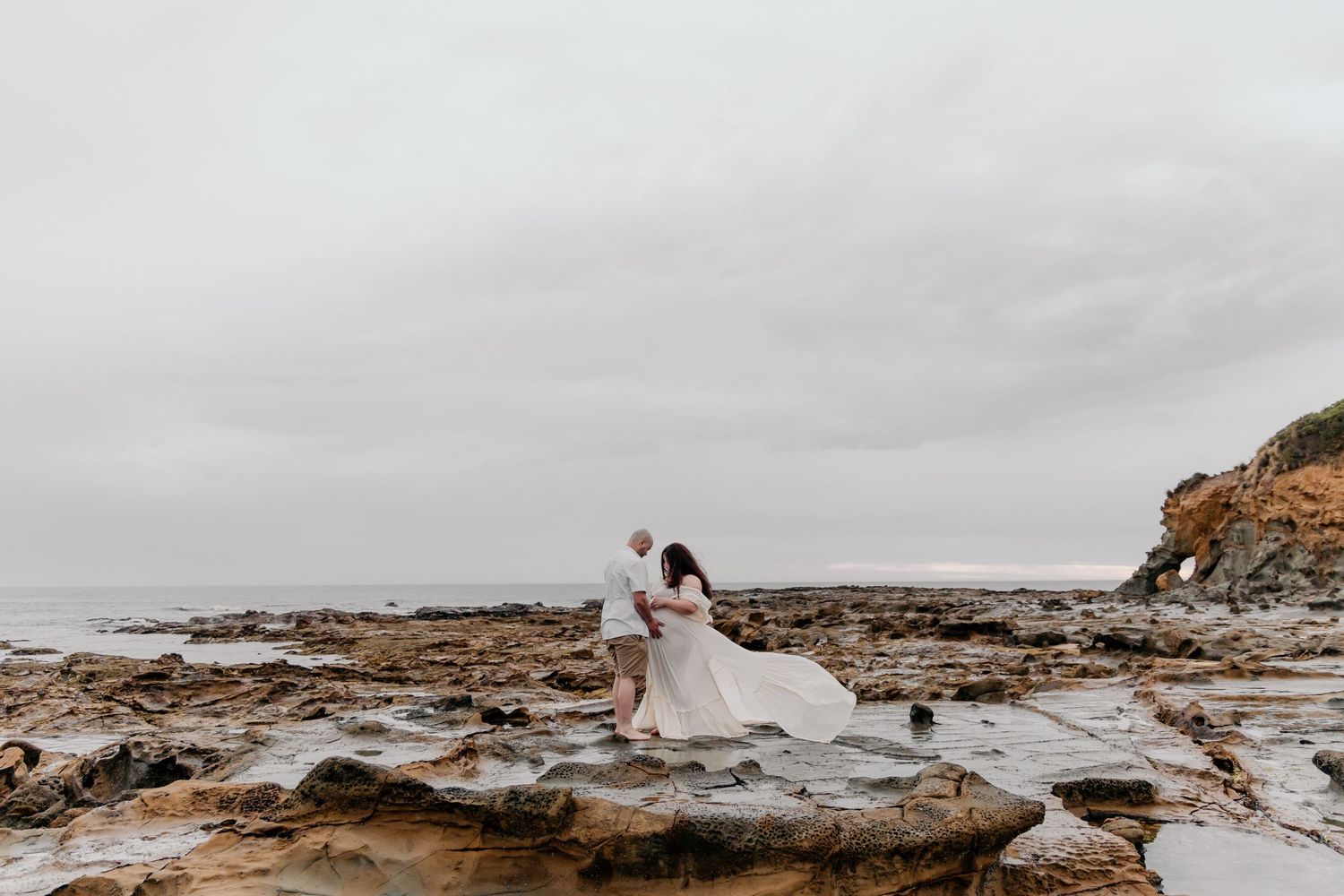 A wedding couple embraces on rocky coastal terrain under an overcast sky.