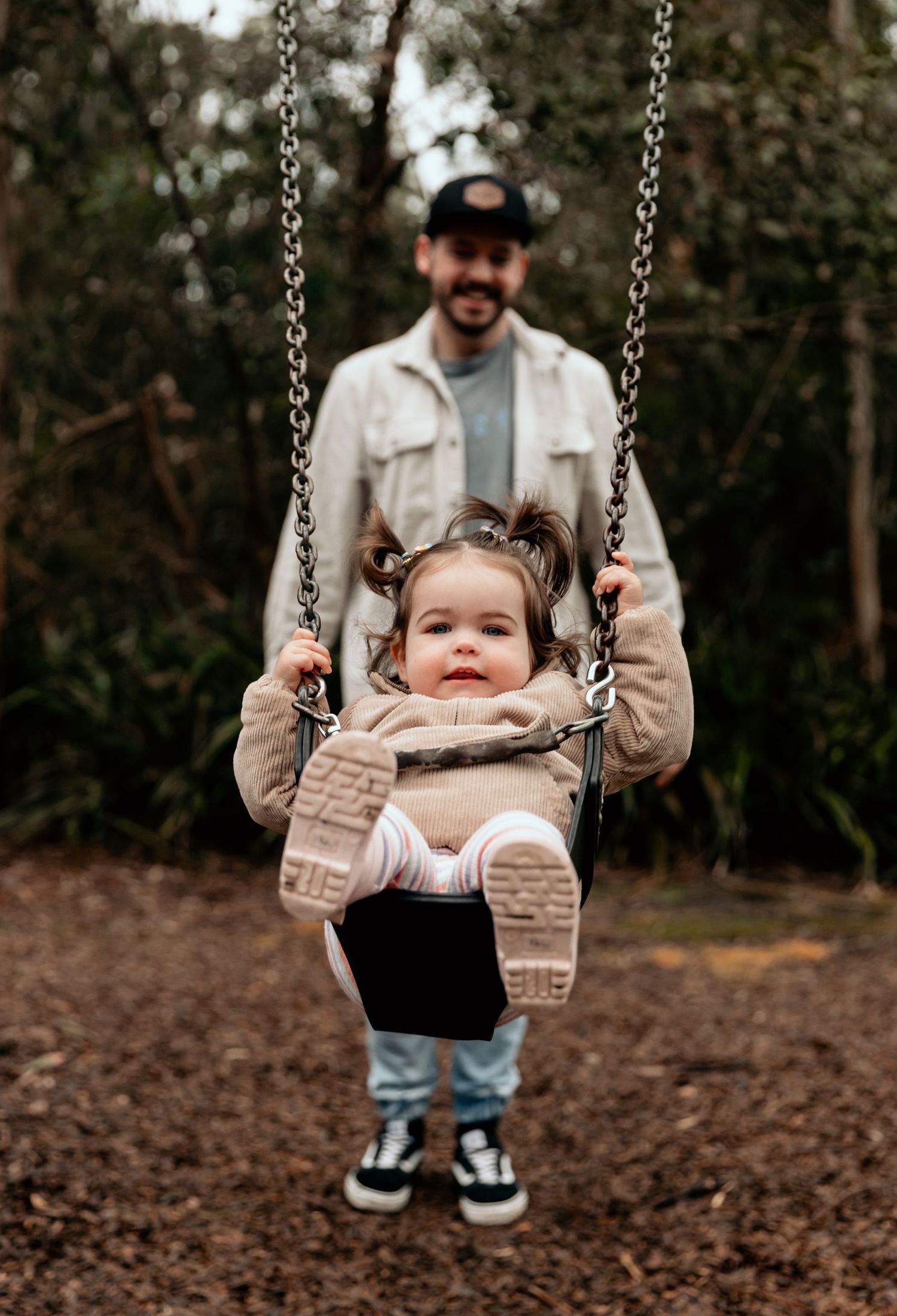 Parent pushing smiling toddler on swing in wooded park setting.