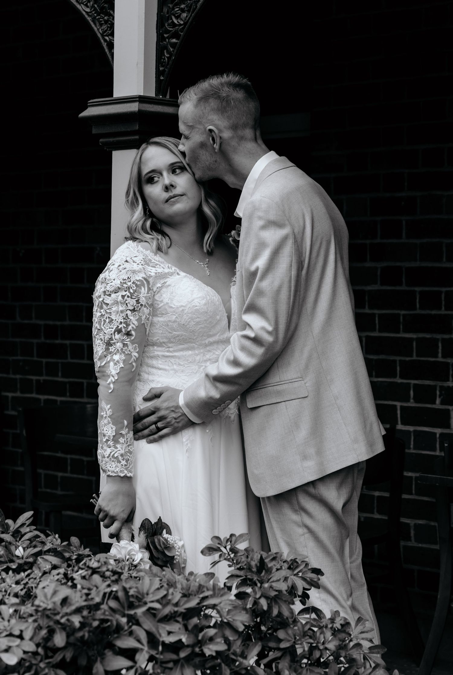 Black and white wedding photo of bride and groom sharing a tender moment, groom kissing bride's temple outdoors.