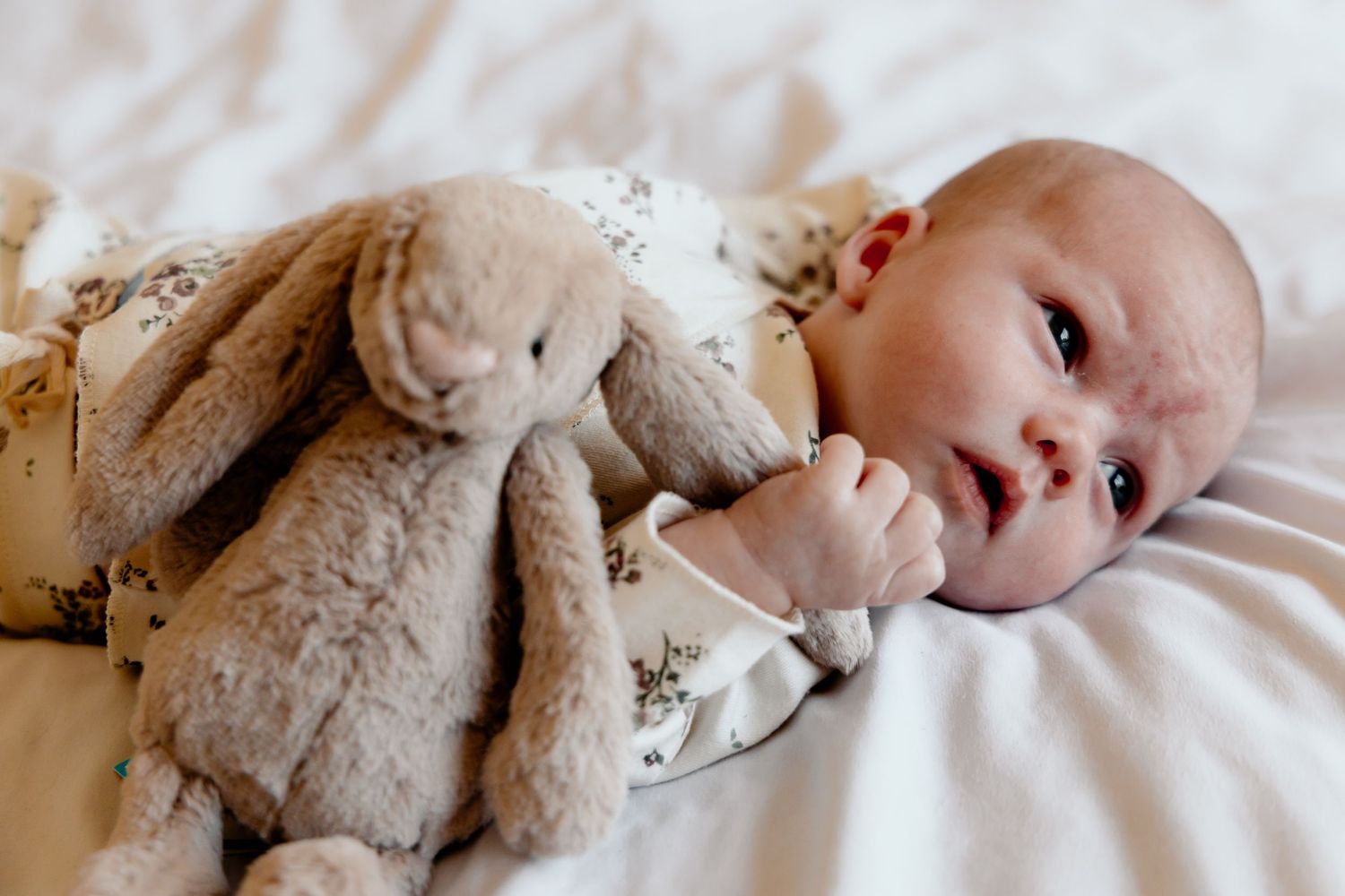 A baby lies peacefully in white bedding while cuddling a soft plush bunny toy.