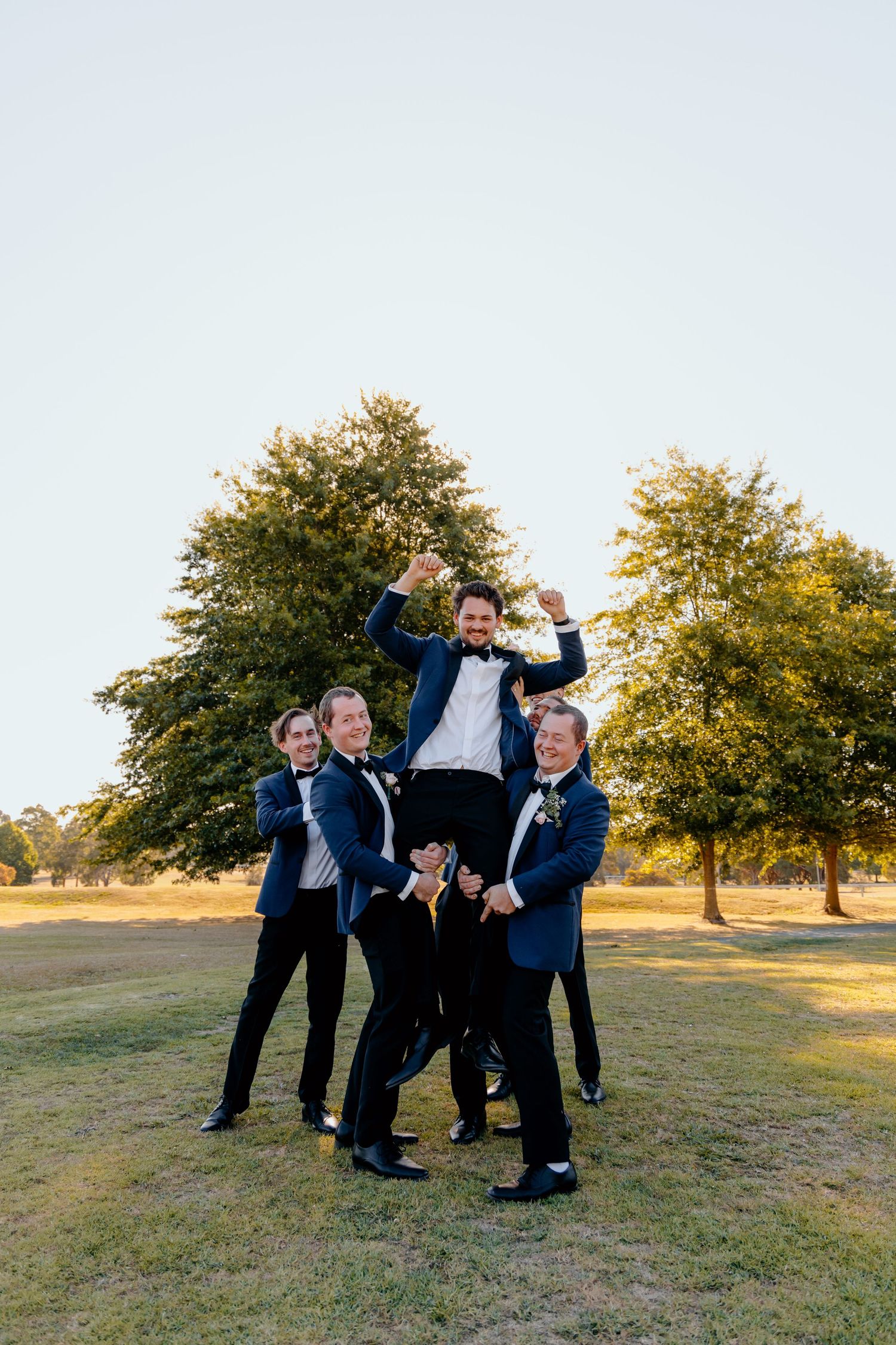 Groomsmen in navy blue suits playfully lift and pose with someone on a grassy field with autumn trees in the background.