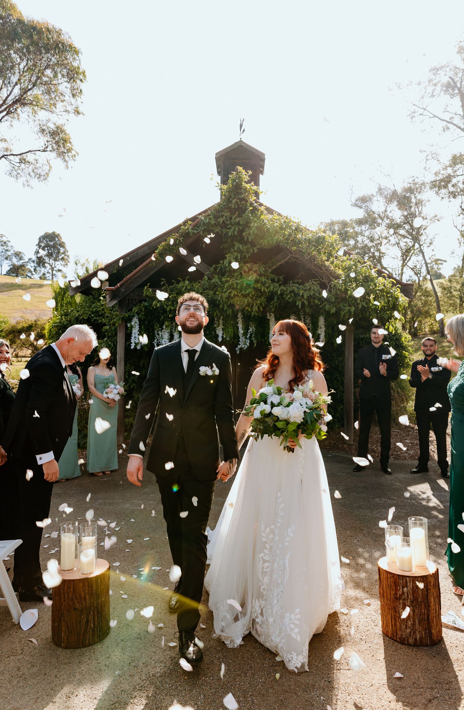 A wedding couple exits a rustic chapel decorated with greenery while guests throw white flower petals in celebration.