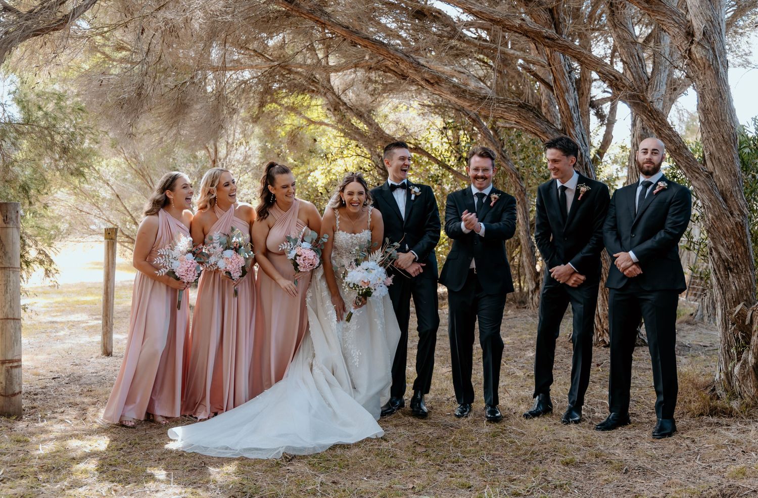Wedding party in pink and black attire poses for a photo under arched tree branches during an outdoor celebration.