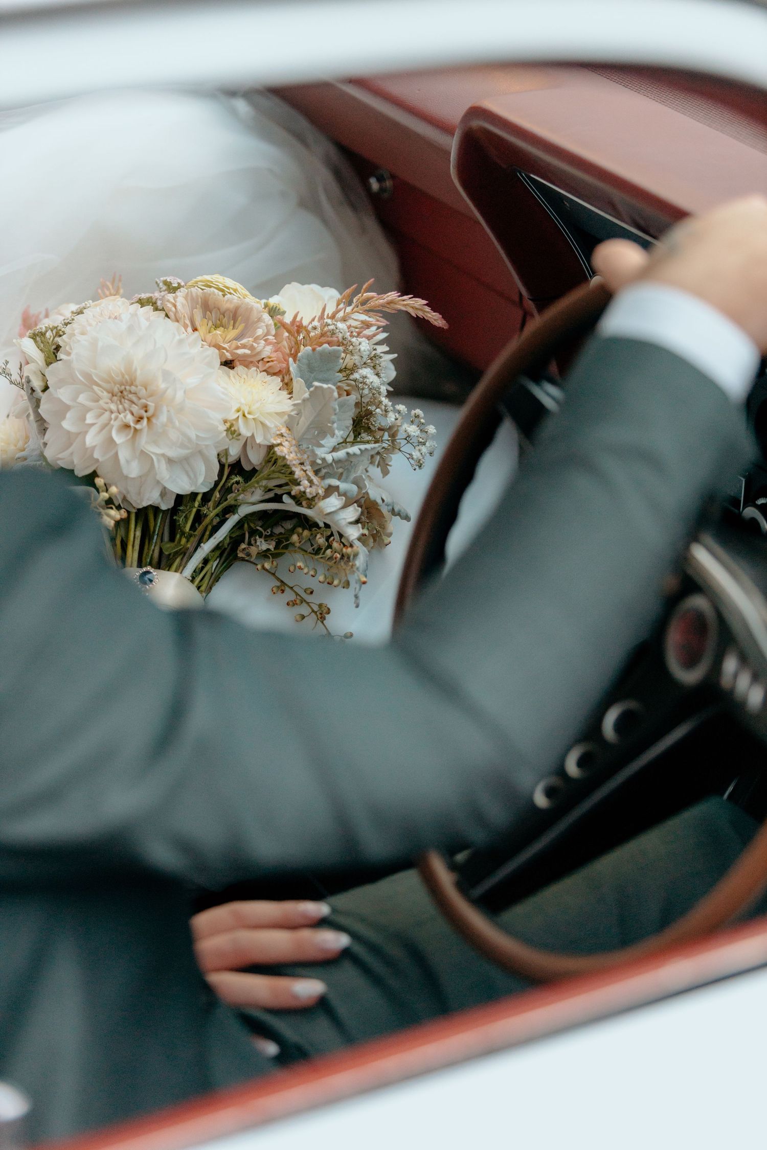 Bride and groom sitting in a vintage car, holding a beautiful white floral bouquet with dahlias on their wedding day.