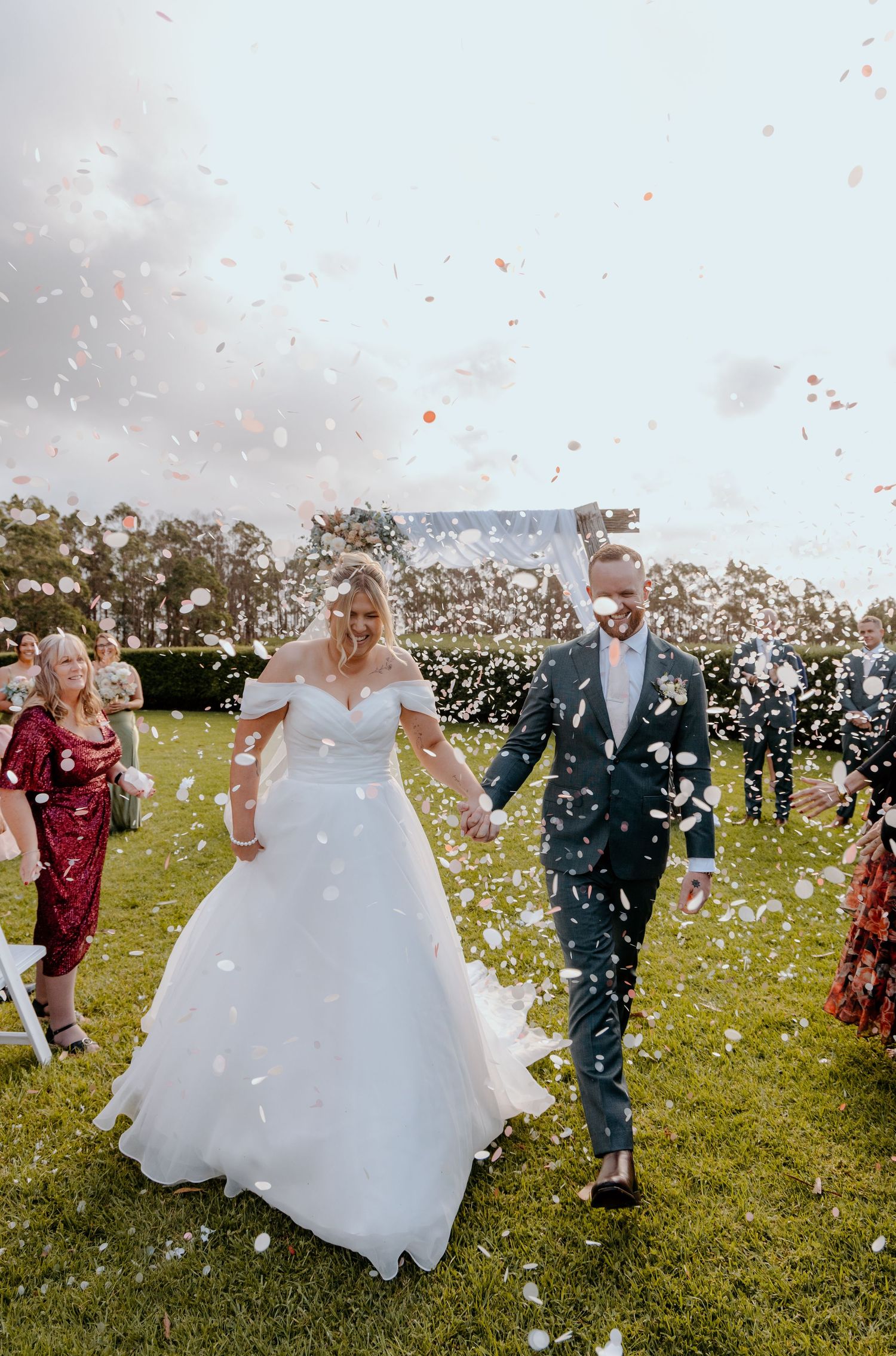 Bride and groom walk through confetti shower on green lawn during outdoor wedding ceremony.