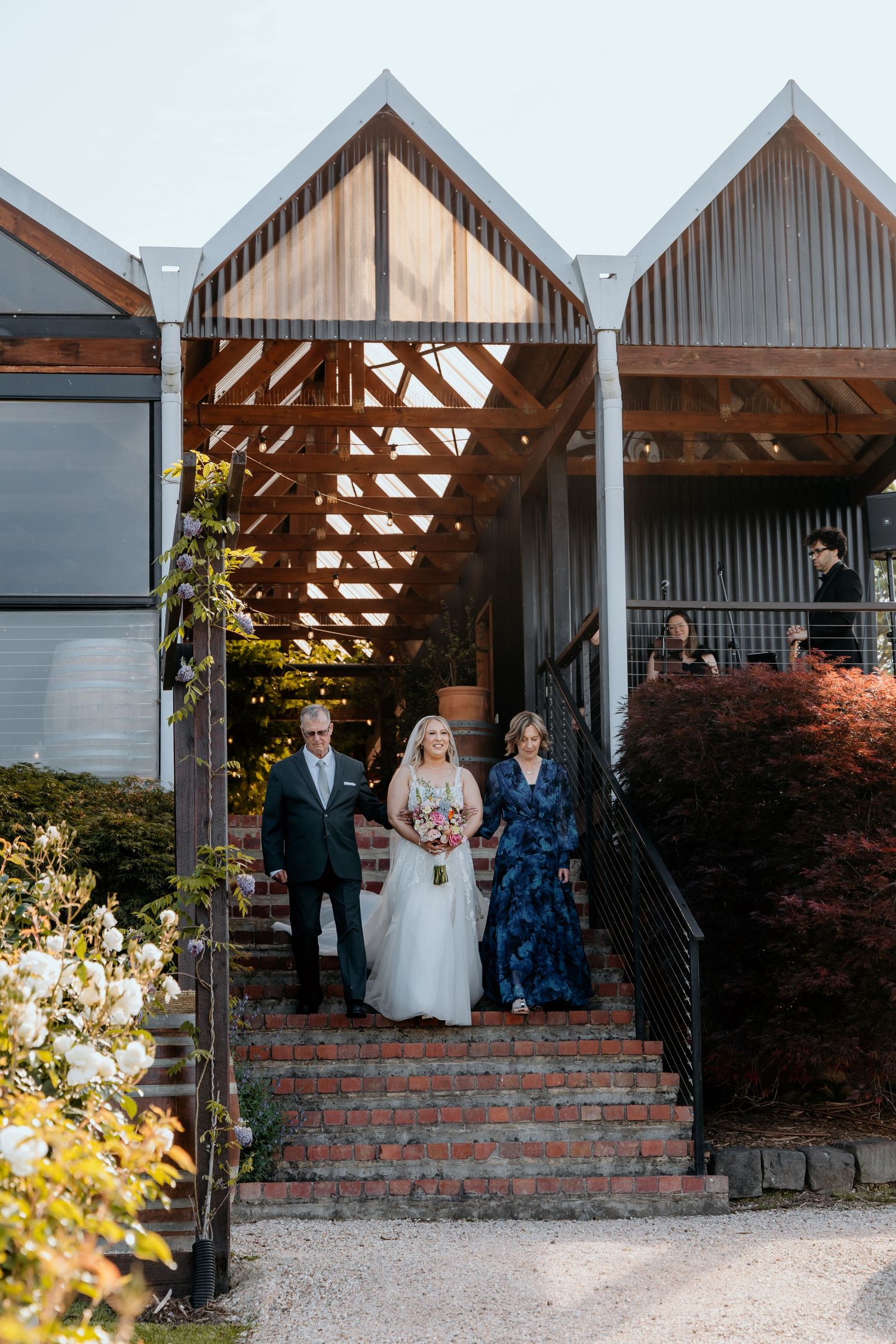 Wedding party stands on rustic brick steps in front of modern white chapel-style venue with exposed beams.