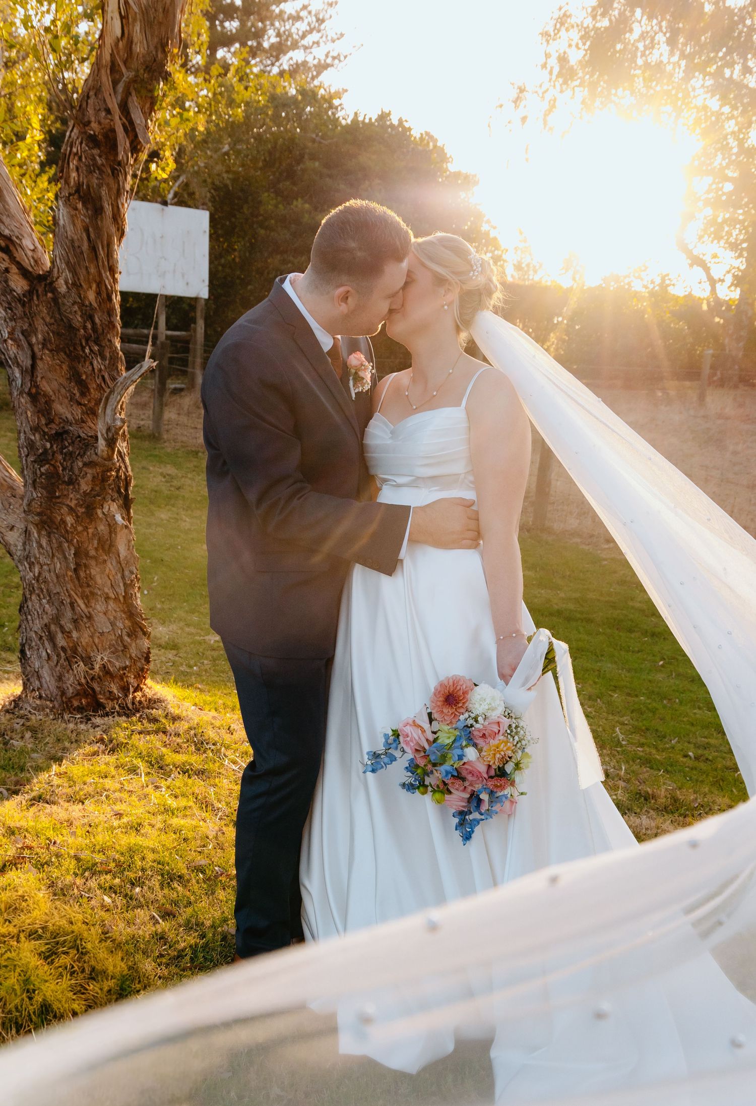 Bride and groom share a golden sunset kiss, bride holding colorful bouquet with veil flowing, at outdoor wedding ceremony.