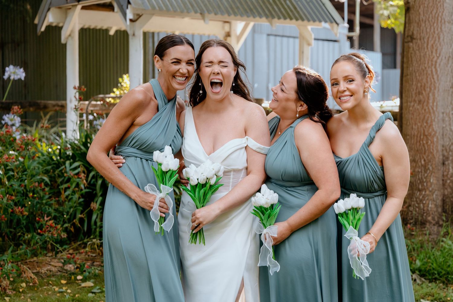 Bride in white gown laughing with three bridesmaids in sage green dresses holding white tulip bouquets outdoors.
