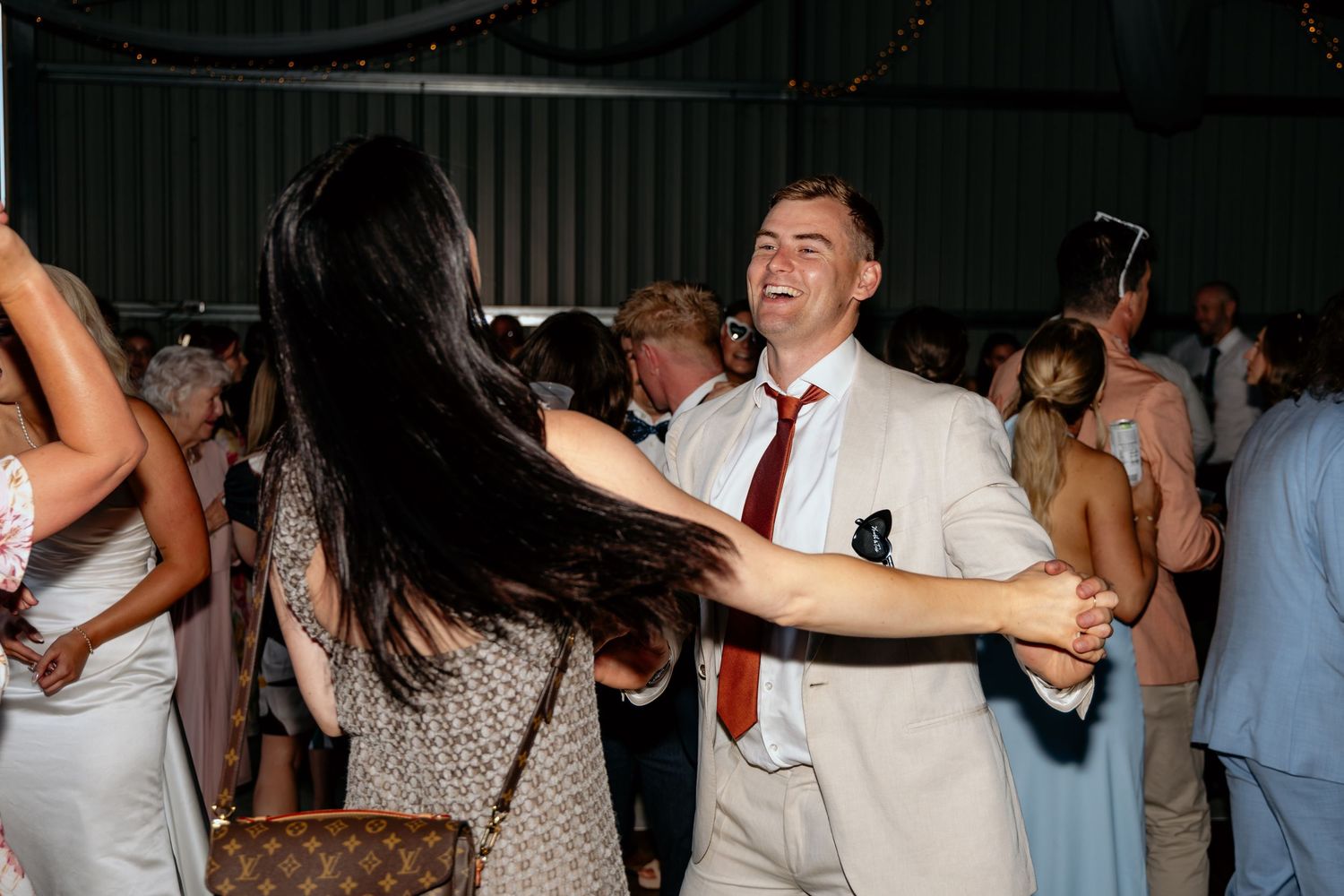 Couple dancing energetically at a wedding reception in a barn venue, man in beige suit with red tie smiling.