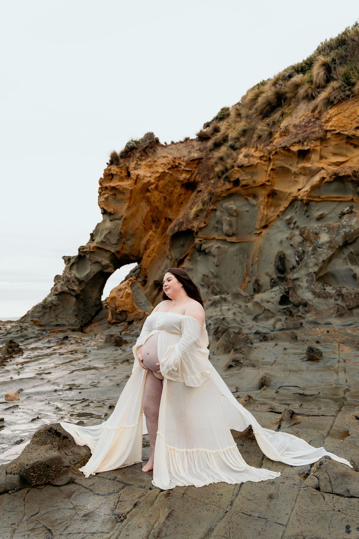 Pregnant woman in flowing cream dress poses by natural rock arch formation at beach.