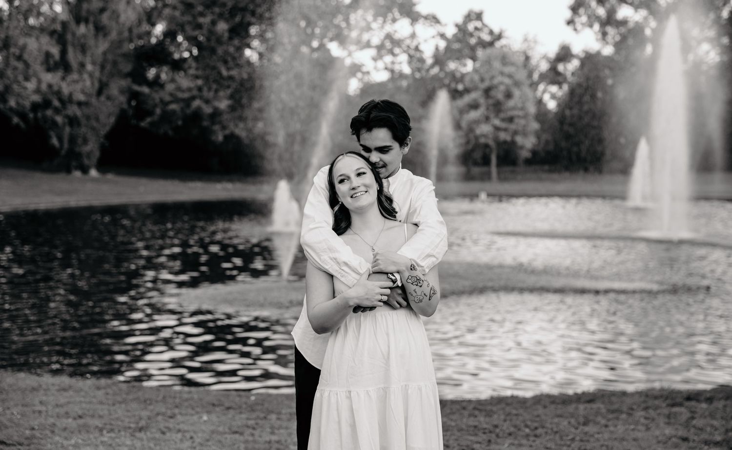 Black and white portrait of couple embracing by fountain in park setting.