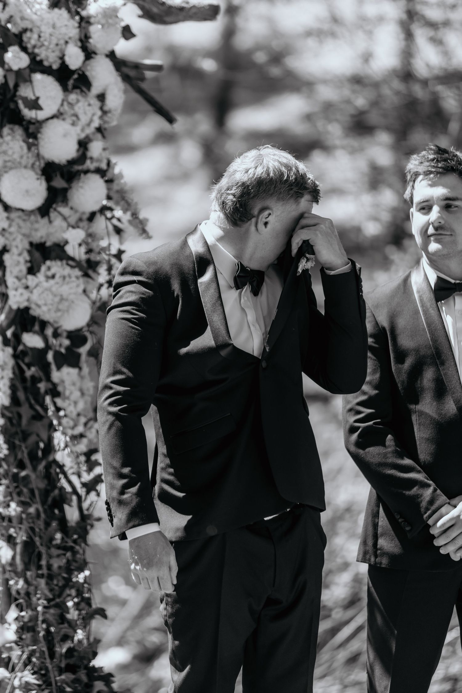 Emotional groom wiping tears at wedding altar in black tuxedo with bow tie, best man standing beside him.