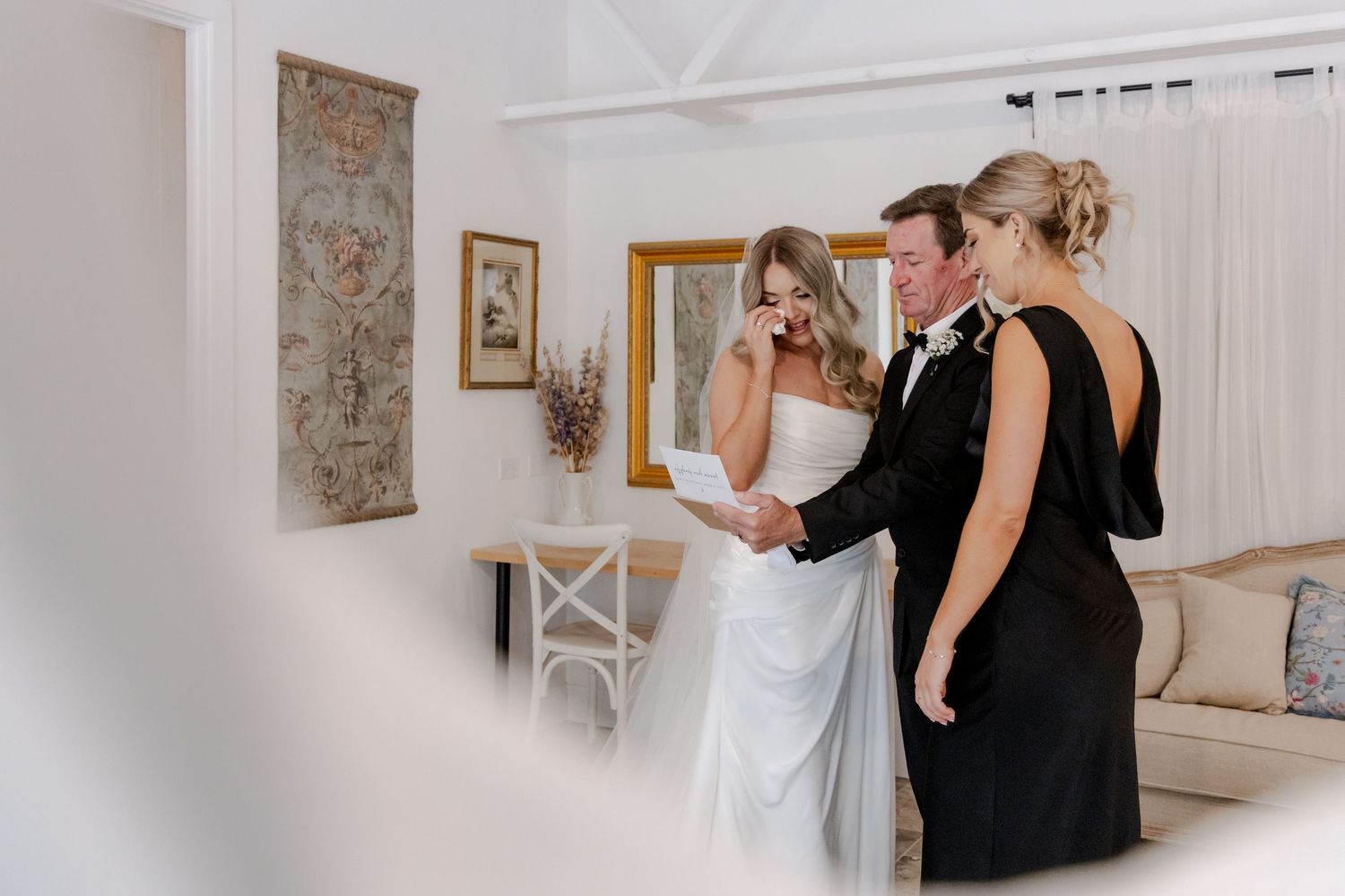 Bride wiping tears during emotional first look moment with groom in elegant white wedding venue interior.