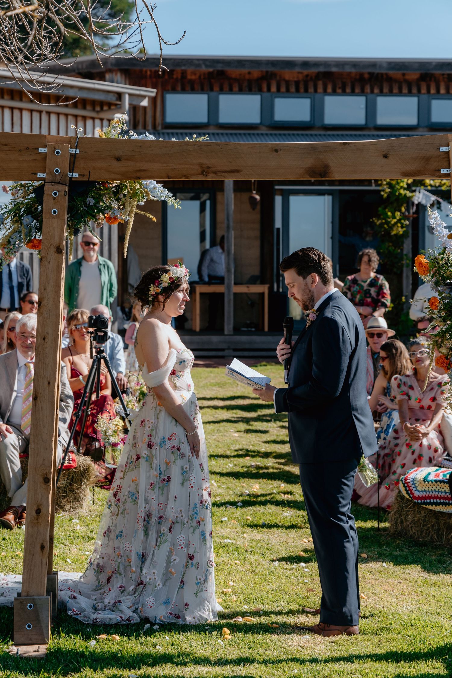Couple exchanging vows outdoors under a wooden arch, bride in floral gown with flower crown, groom in navy suit.