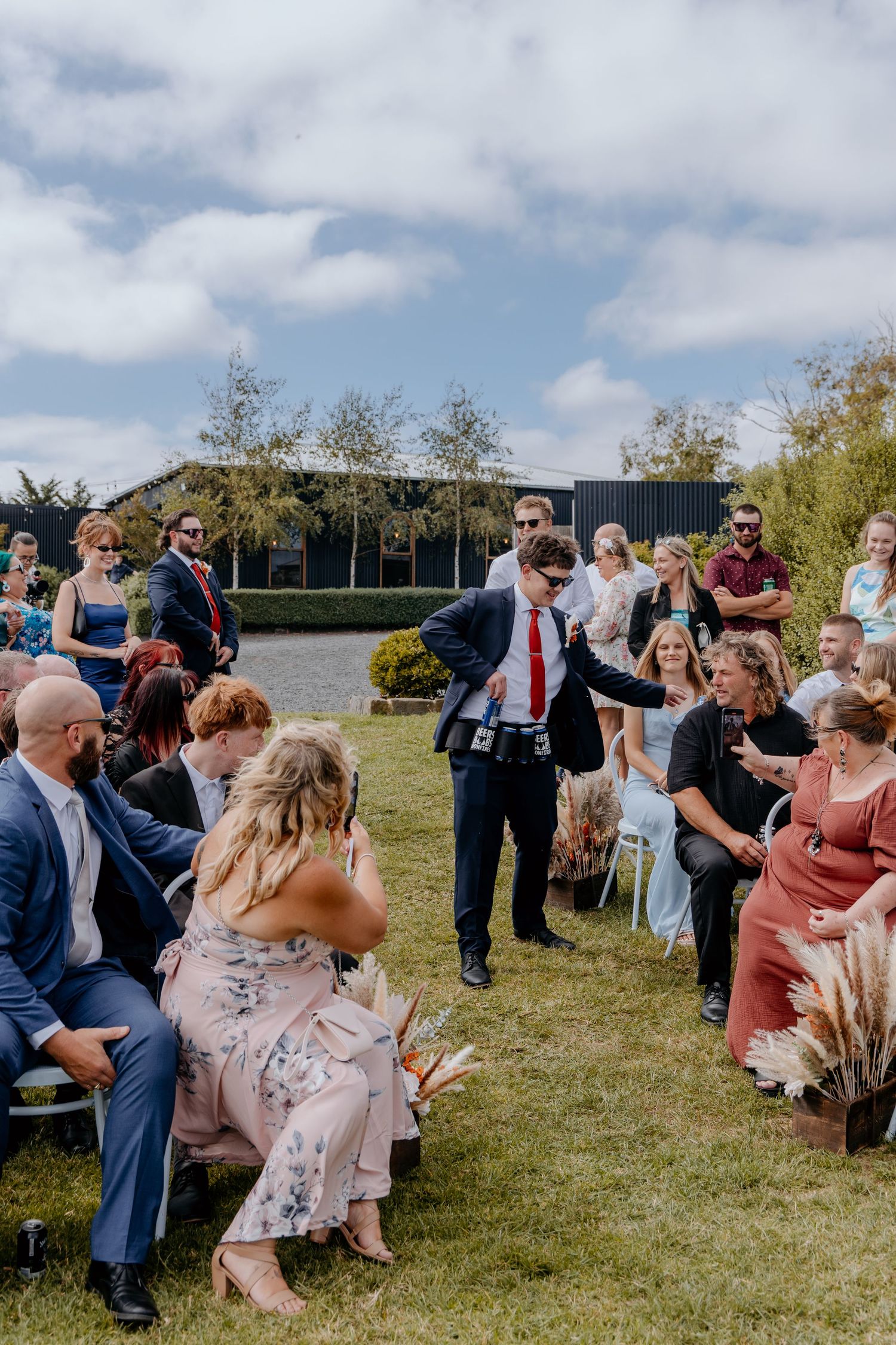 Groomsman in navy suit and red tie interacts with seated wedding guests during outdoor ceremony.