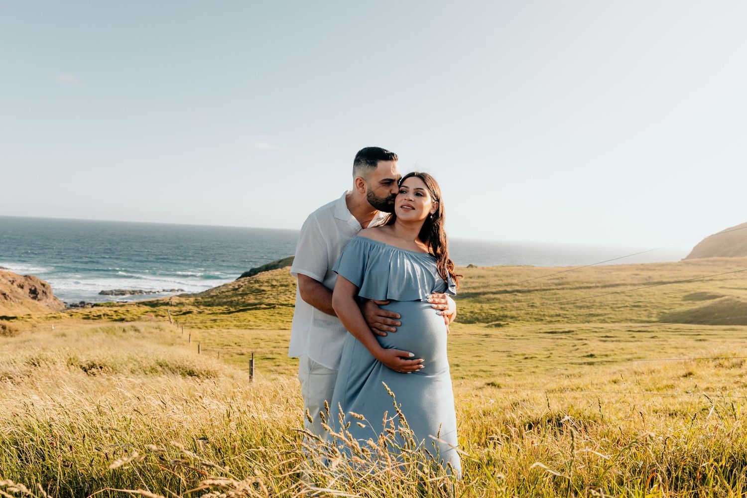 Expecting couple embracing on coastal clifftop with ocean views in background.