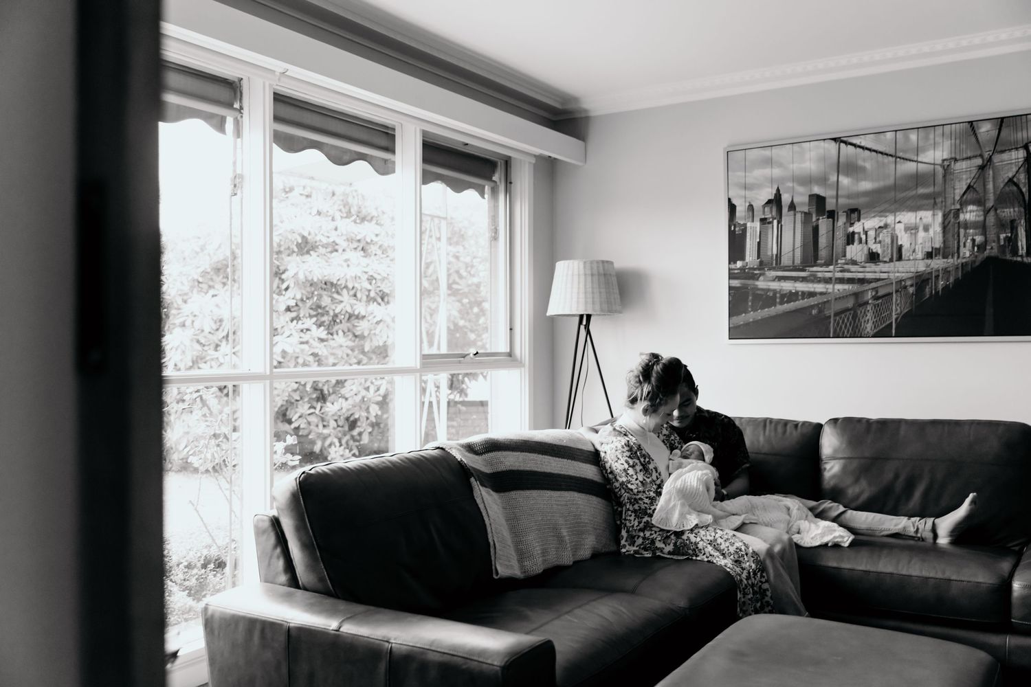 A person relaxes on a grey couch in a modern living room with large windows and city artwork on the wall.