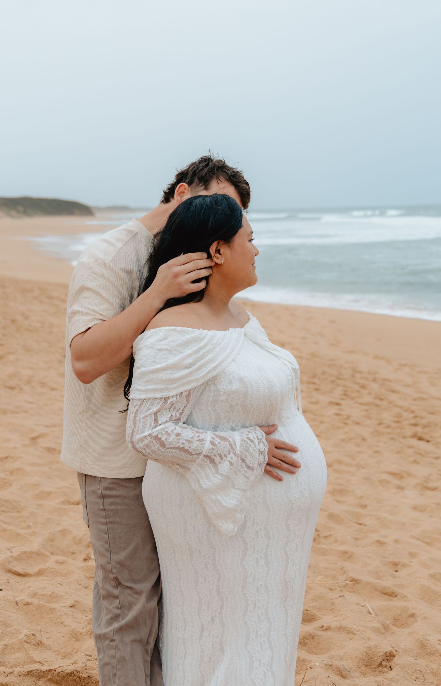 Couple shares intimate moment on sandy beach as waves crash in background, wearing white dress and beige outfit.