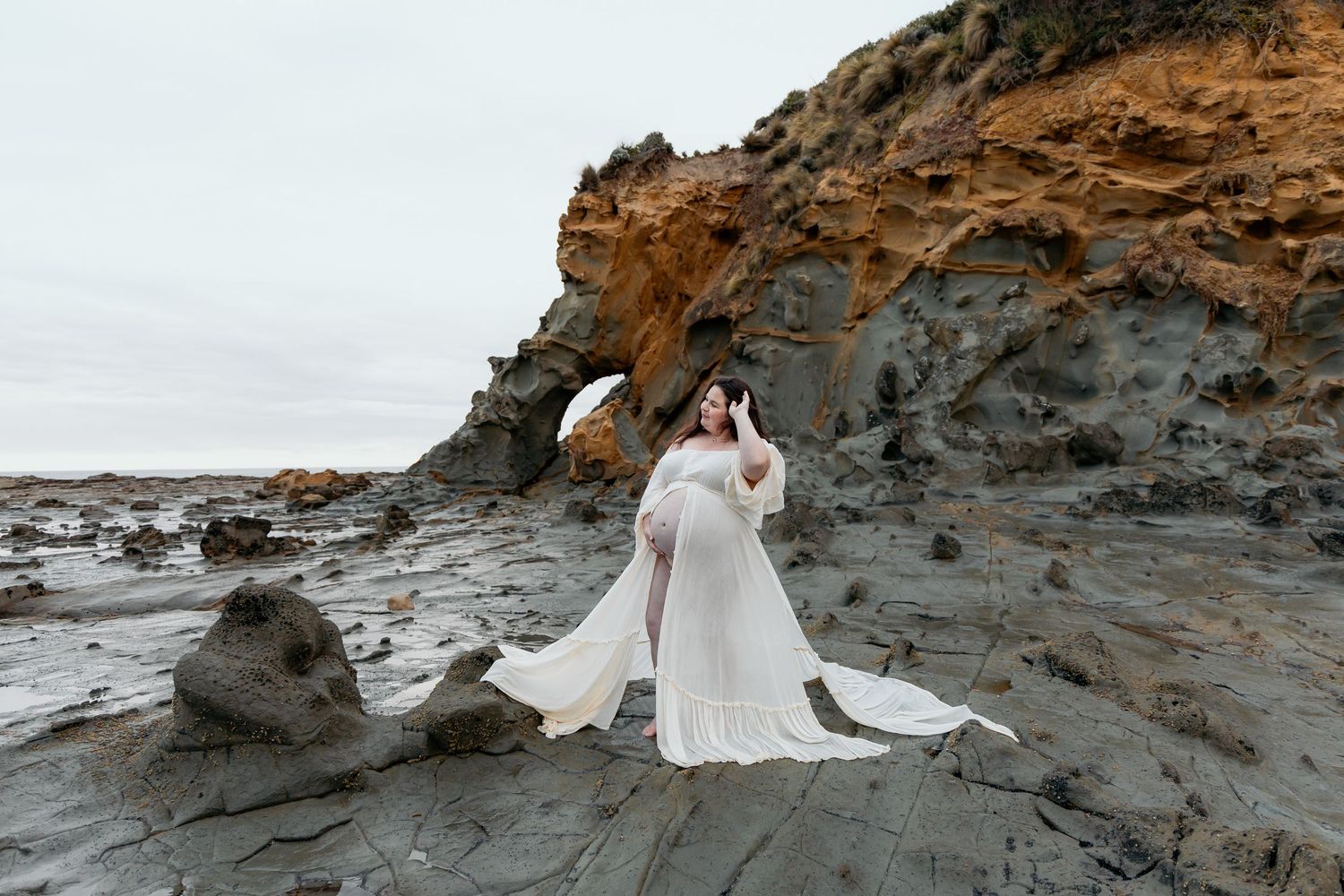 A figure in a flowing white gown stands on dramatic coastal rocks against a rugged cliff backdrop.