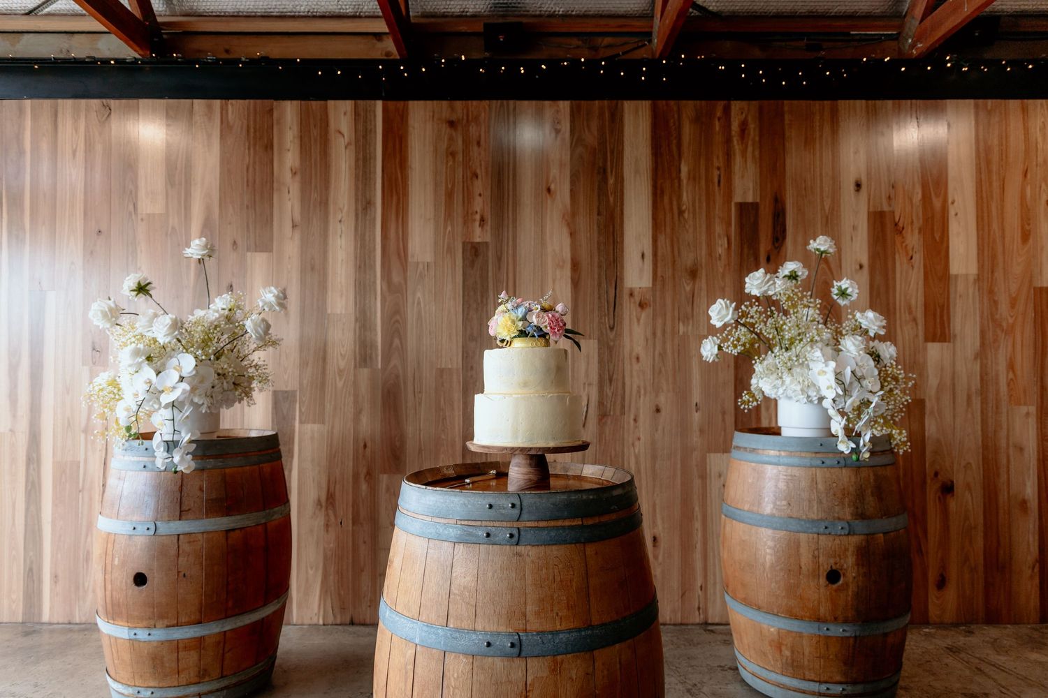 A rustic wedding cake displayed on wooden wine barrels with white floral arrangements in a barn-style venue.