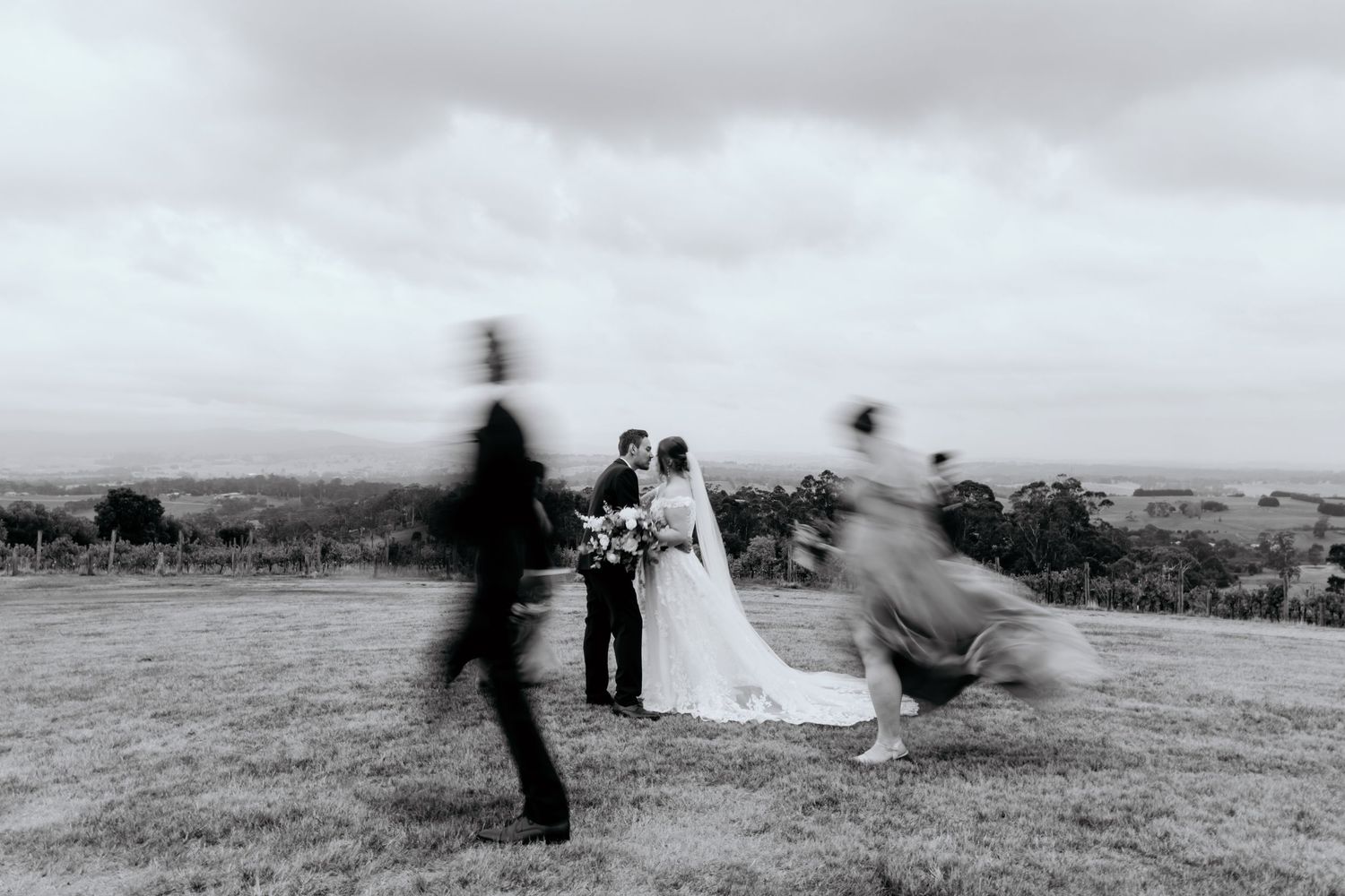 Black and white artistic motion blur photograph showing a couple in wedding attire on a hilltop with blurred figures running past.