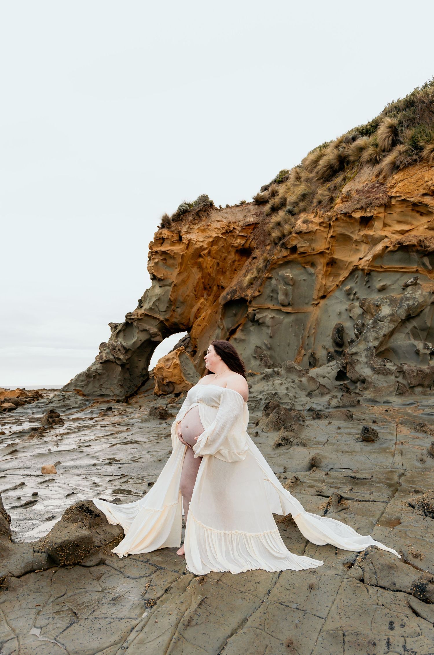 A figure in a flowing white dress poses gracefully on rugged coastal rock formations at sunset.