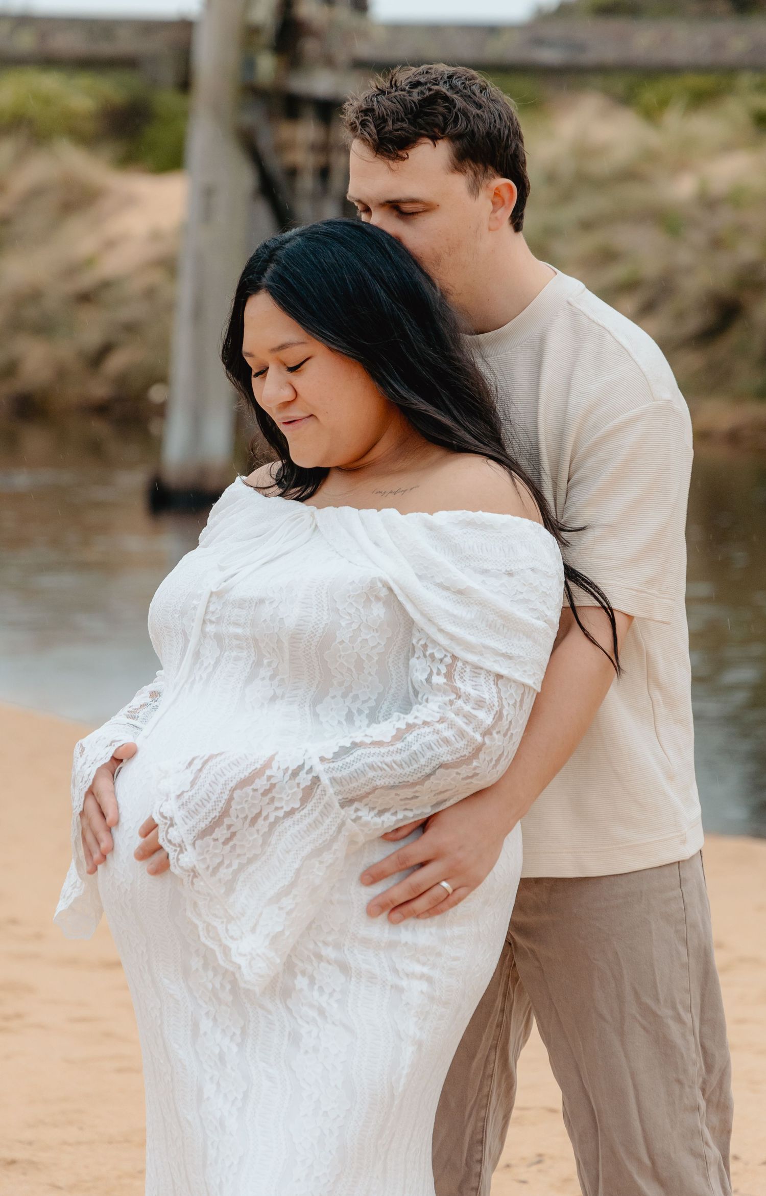 A romantic couple embraces on a sandy beach, with the person in white being dipped during their intimate photoshoot.