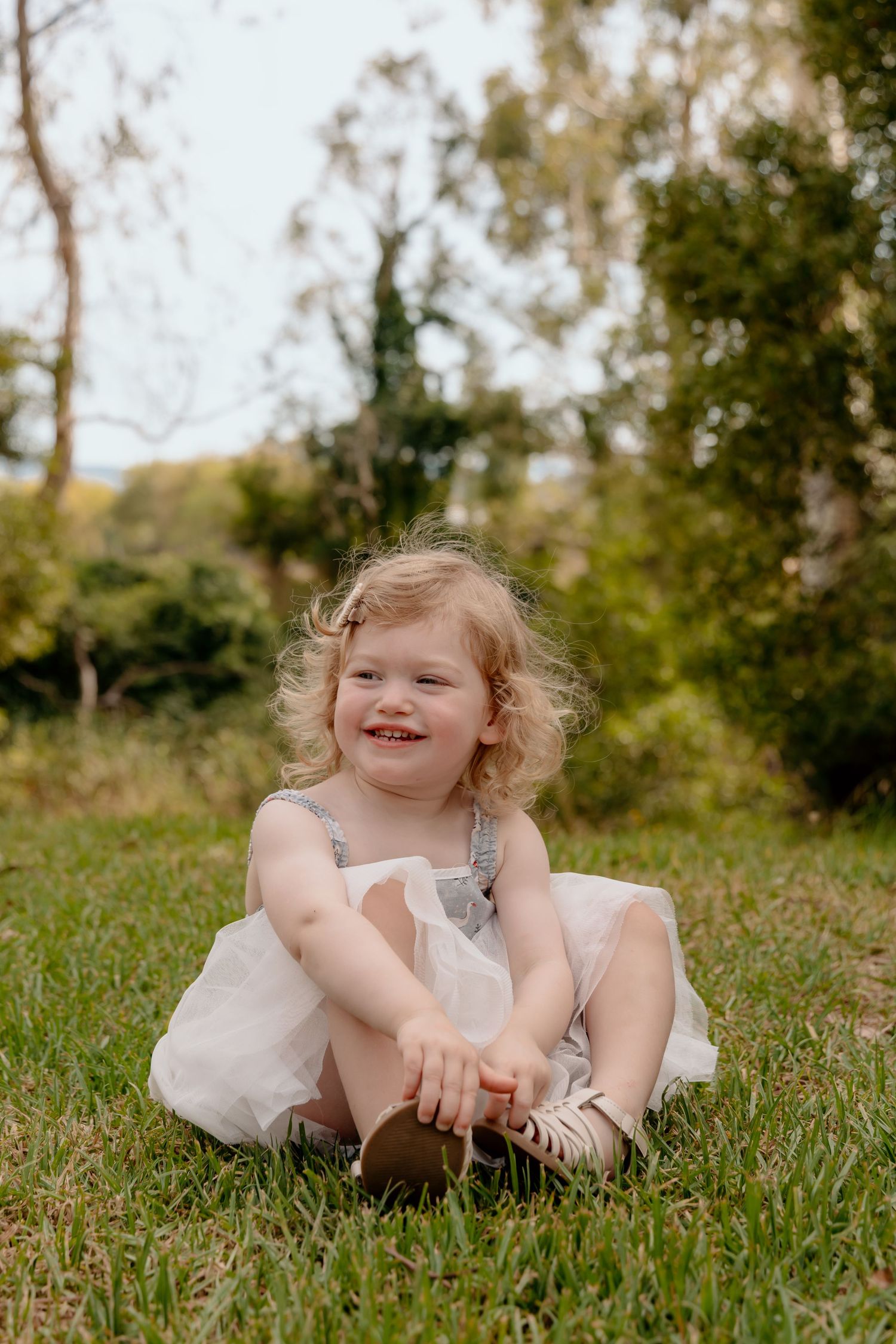 Child in dress sits laughing in grassy field on sunny day.