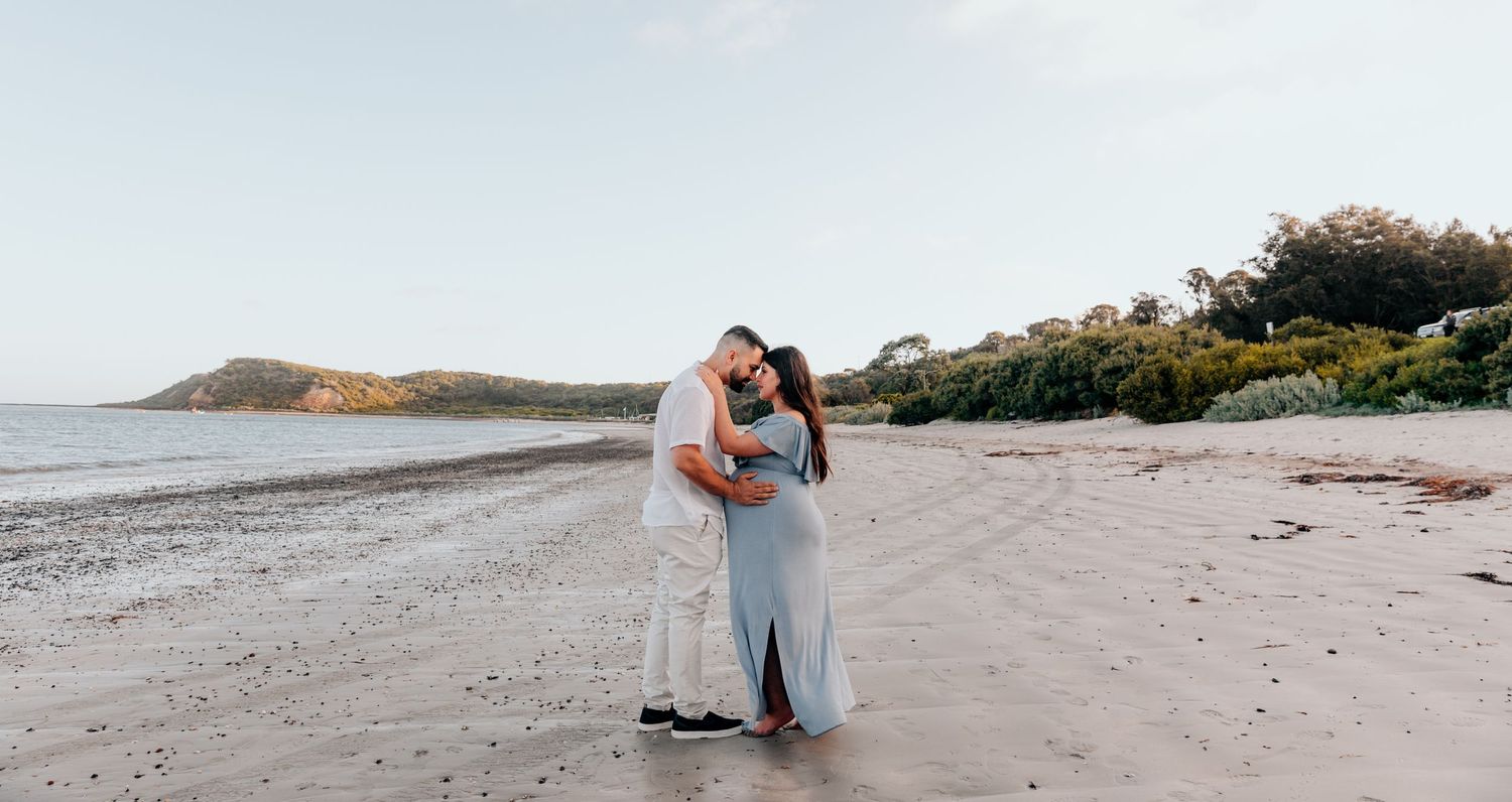 Maternity photo of couple embracing on sandy beach with coastal cliffs in background.