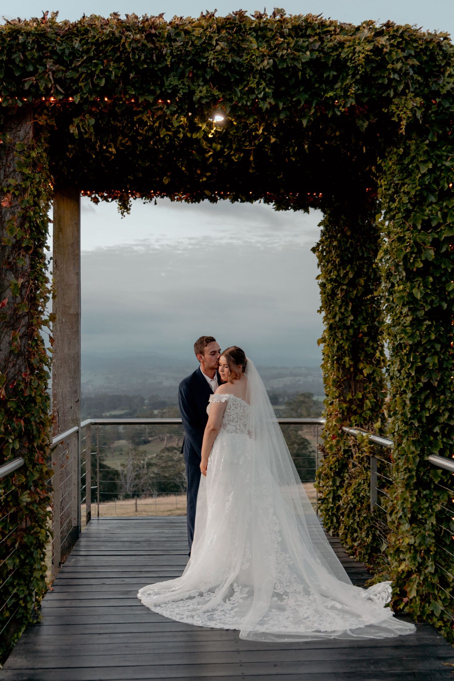 A romantic moment captured at a scenic overlook with a vine-covered arch and dramatic mountain vista in the background.