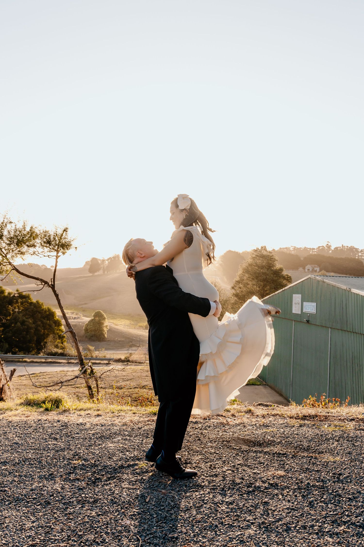 A couple dances together at sunset in front of a rustic barn with sunlit hills in the background.